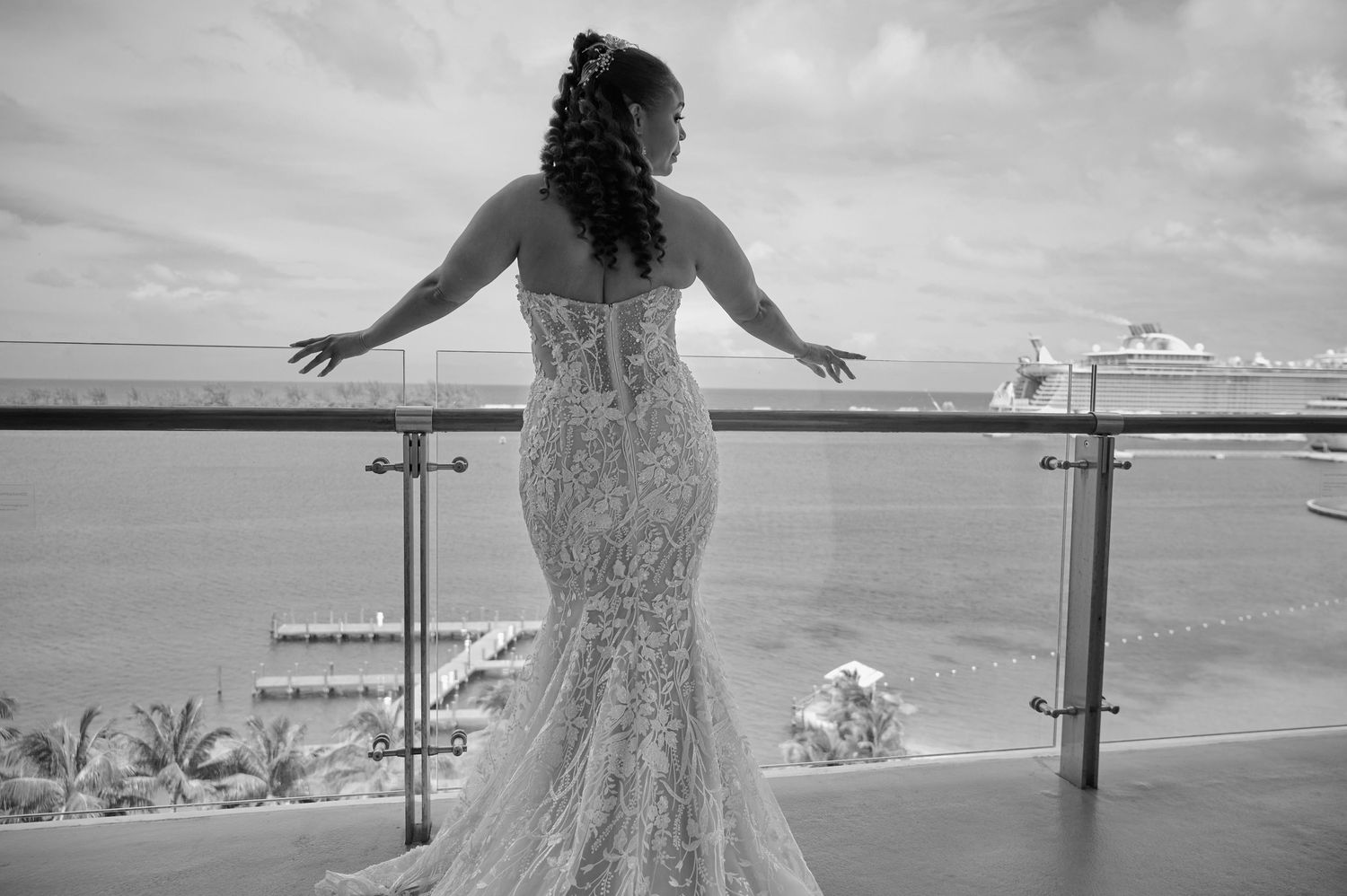 Bride in fitted lace mermaid gown stands on balcony overlooking ocean in black and white photo.