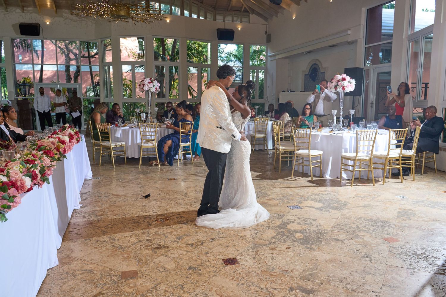 Newlyweds share their first dance in an elegant open-air reception hall with guests and floral arrangements.