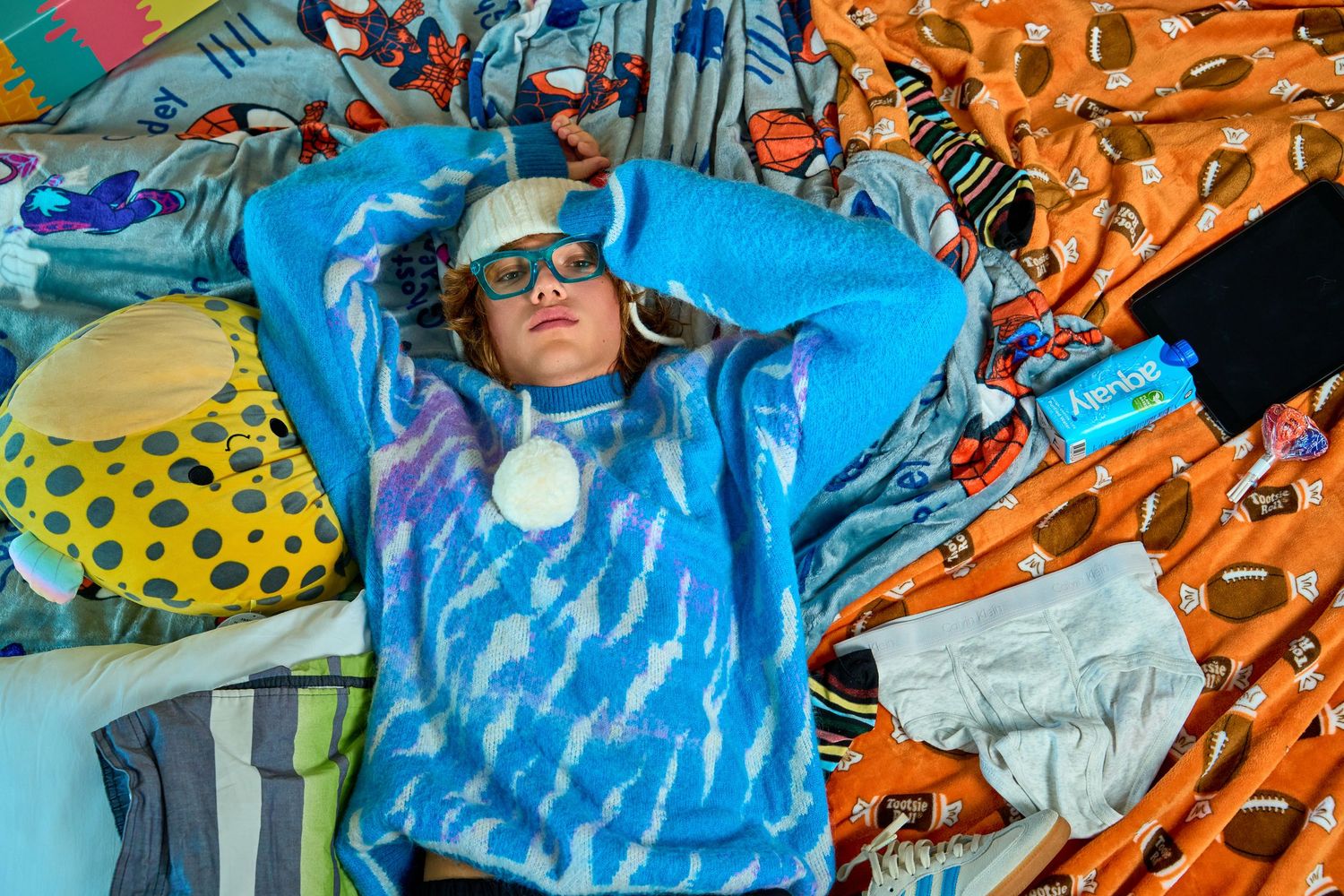 Young person in blue patterned sweatshirt and cap lies relaxed among colorful patterned fabrics and objects.