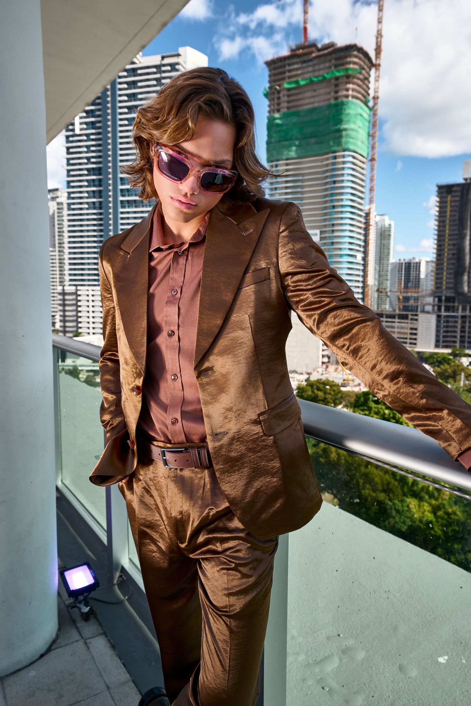 Stylish man in metallic bronze suit and tinted sunglasses poses on urban balcony with city skyline backdrop.