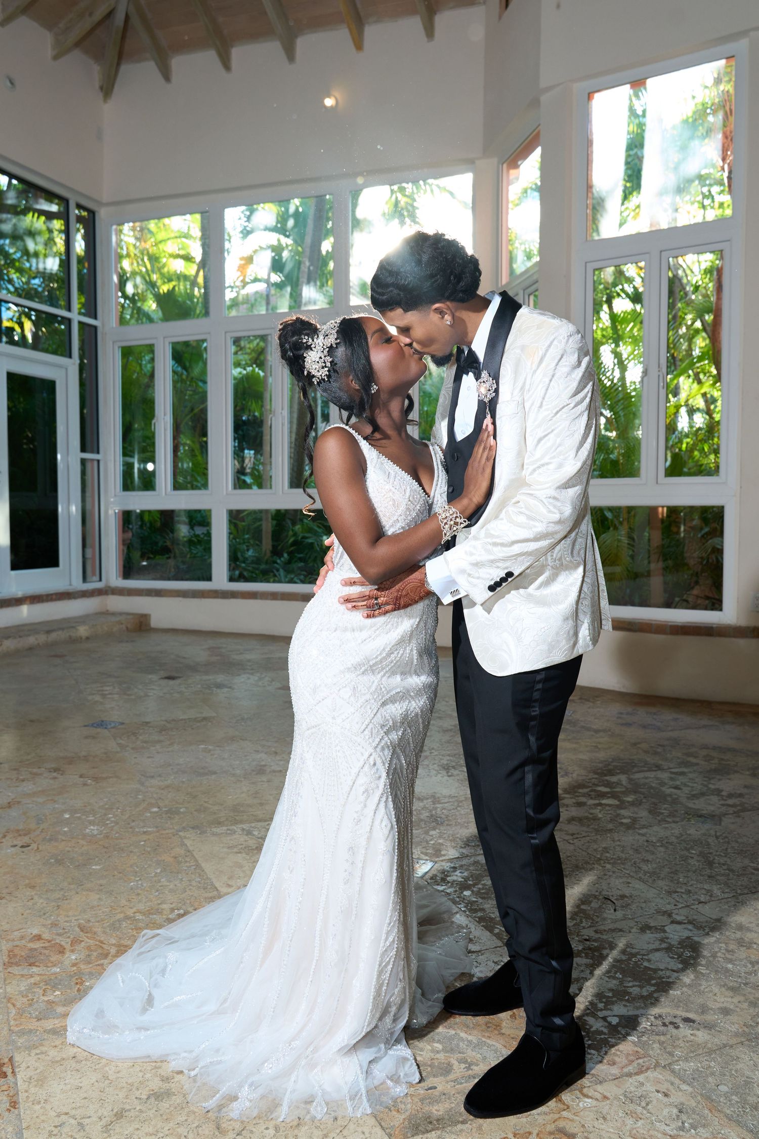 Bride and groom sharing a kiss in elegant sunlit venue with large windows and tropical views.