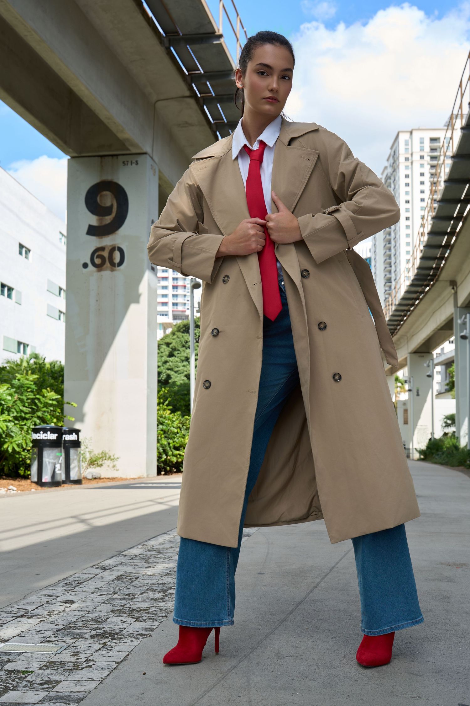 Confident woman in beige trench coat, red tie, jeans, and red heels poses under city overpass structure.