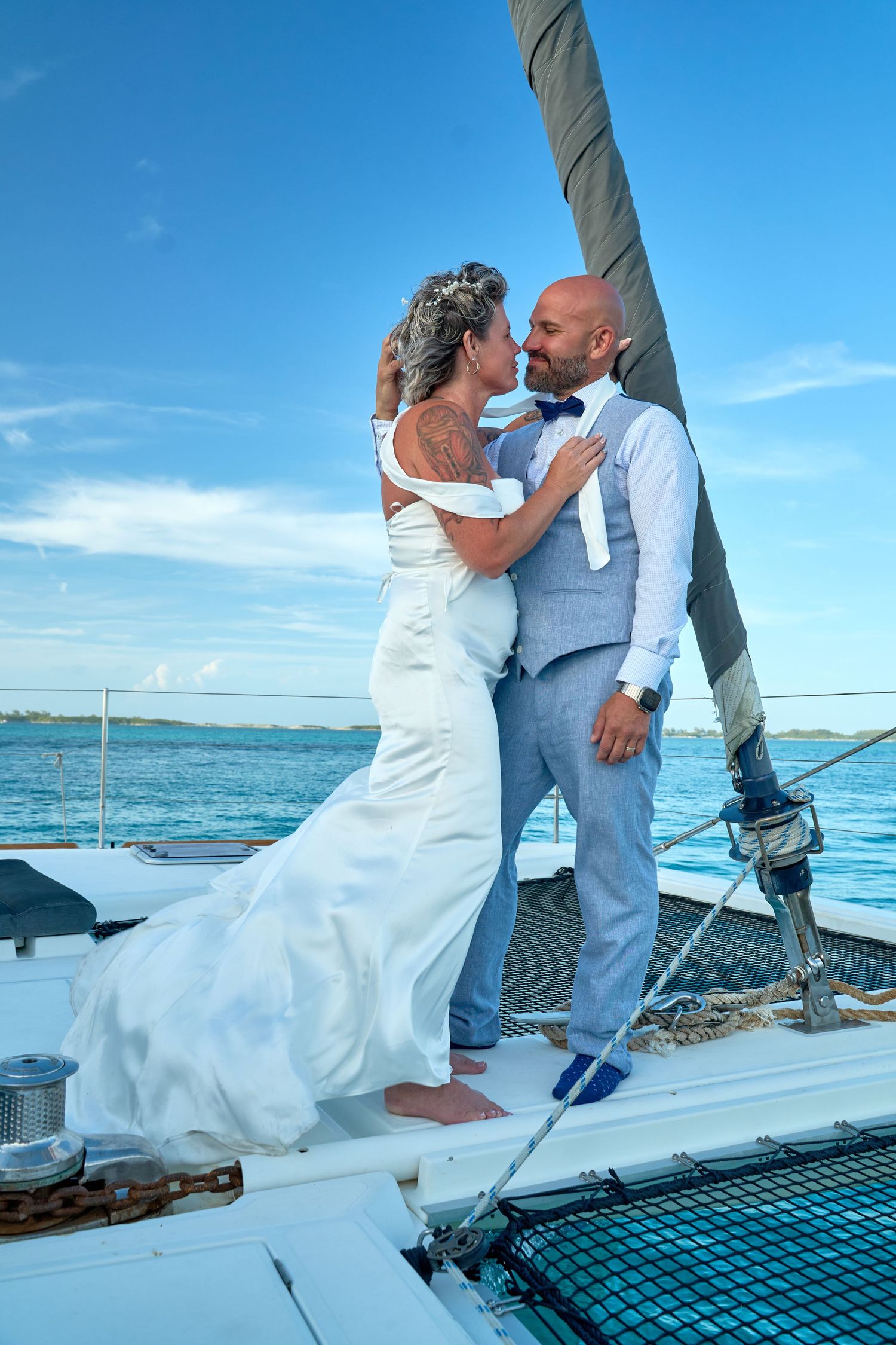 Bride and groom embrace on a sailboat deck with blue ocean and sky backdrop on their wedding day.