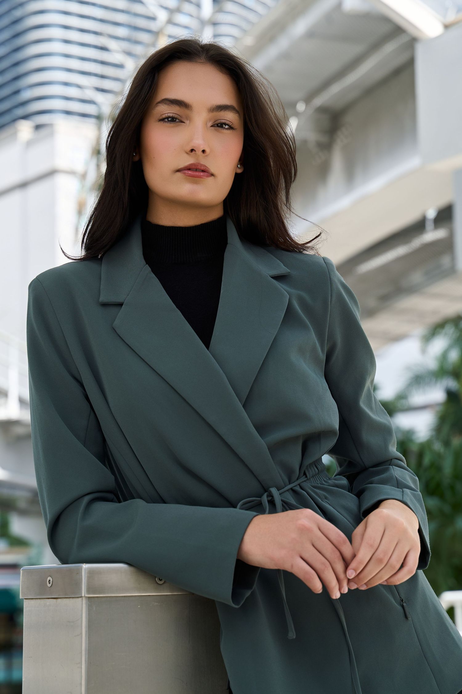 Sophisticated woman in teal belted blazer leans against railing in modern urban outdoor setting.