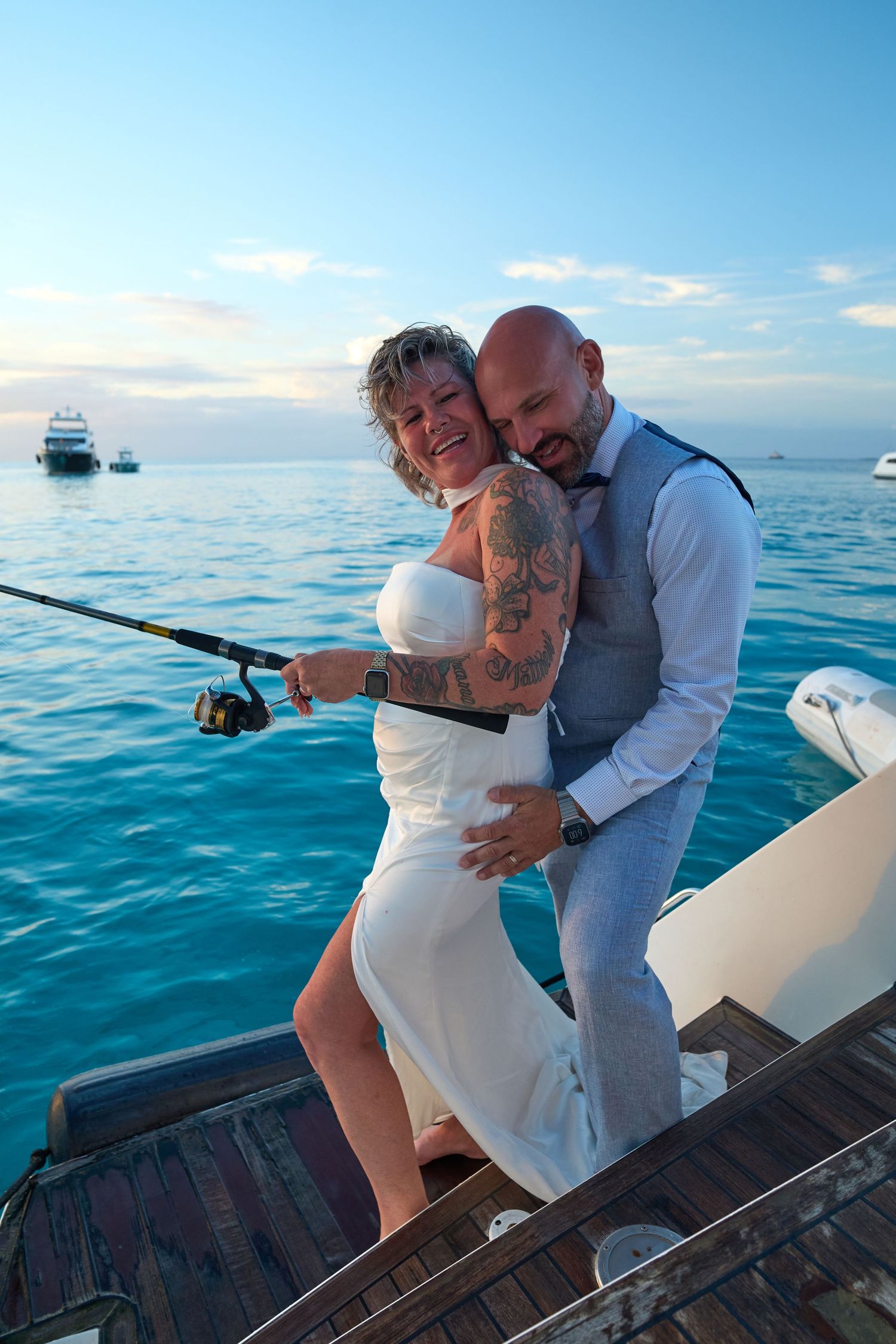 Tattooed bride holding fishing rod embraced by bald groom on boat deck at ocean sunset.