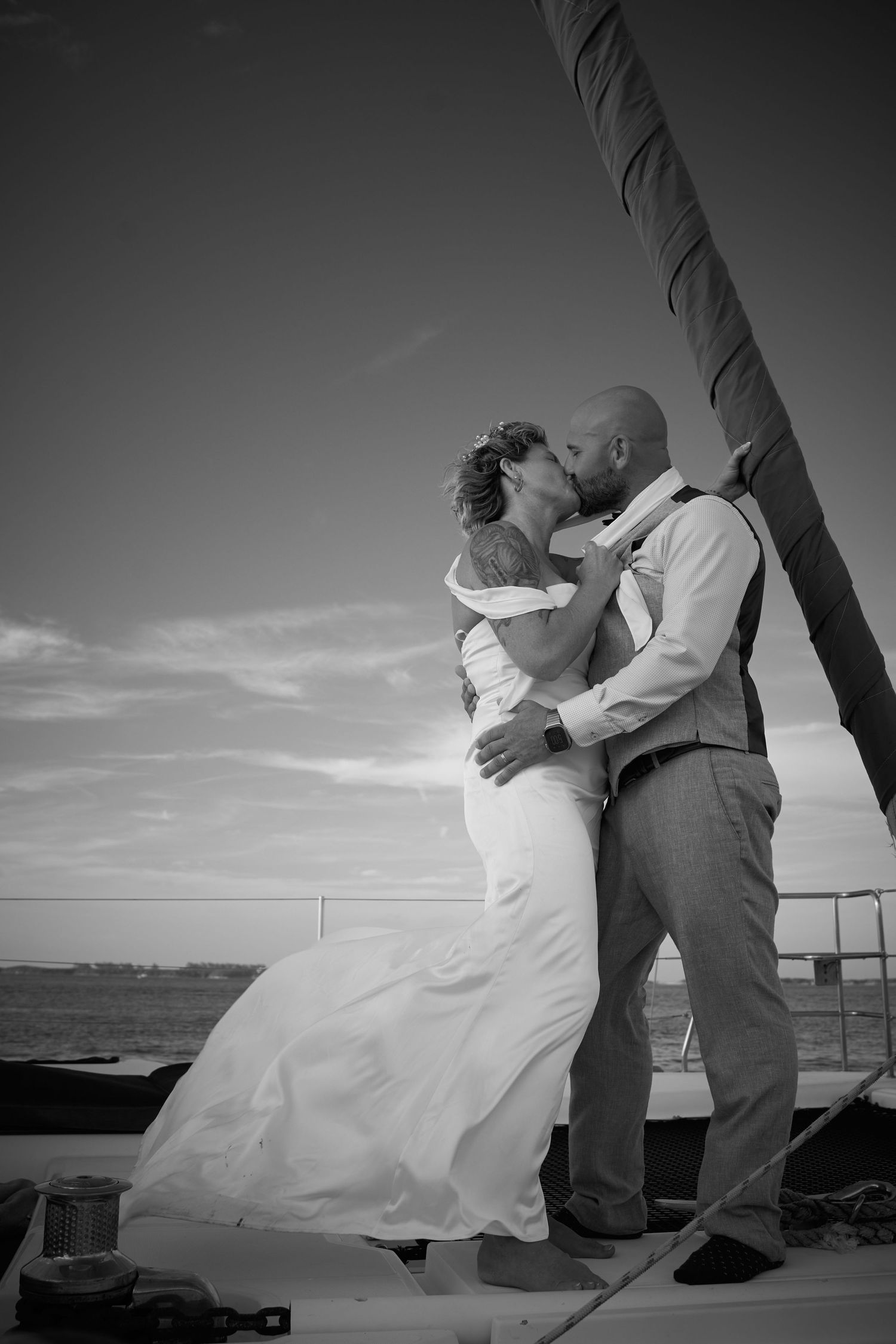 Black and white romantic photo of bride and groom kissing on a sailboat with ocean backdrop.