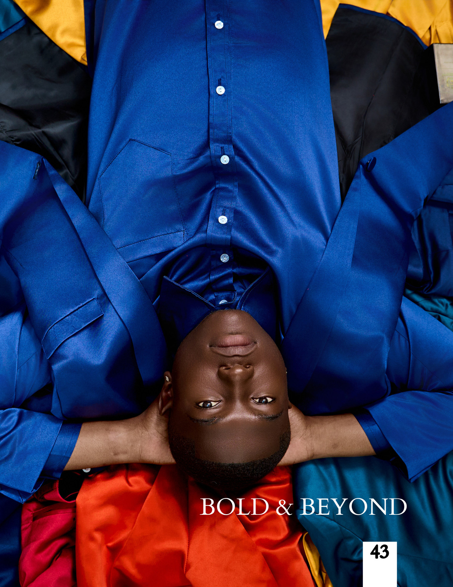 Man in bold blue shirt lying down surrounded by colorful fabric for Bold and Beyond magazine page 43.