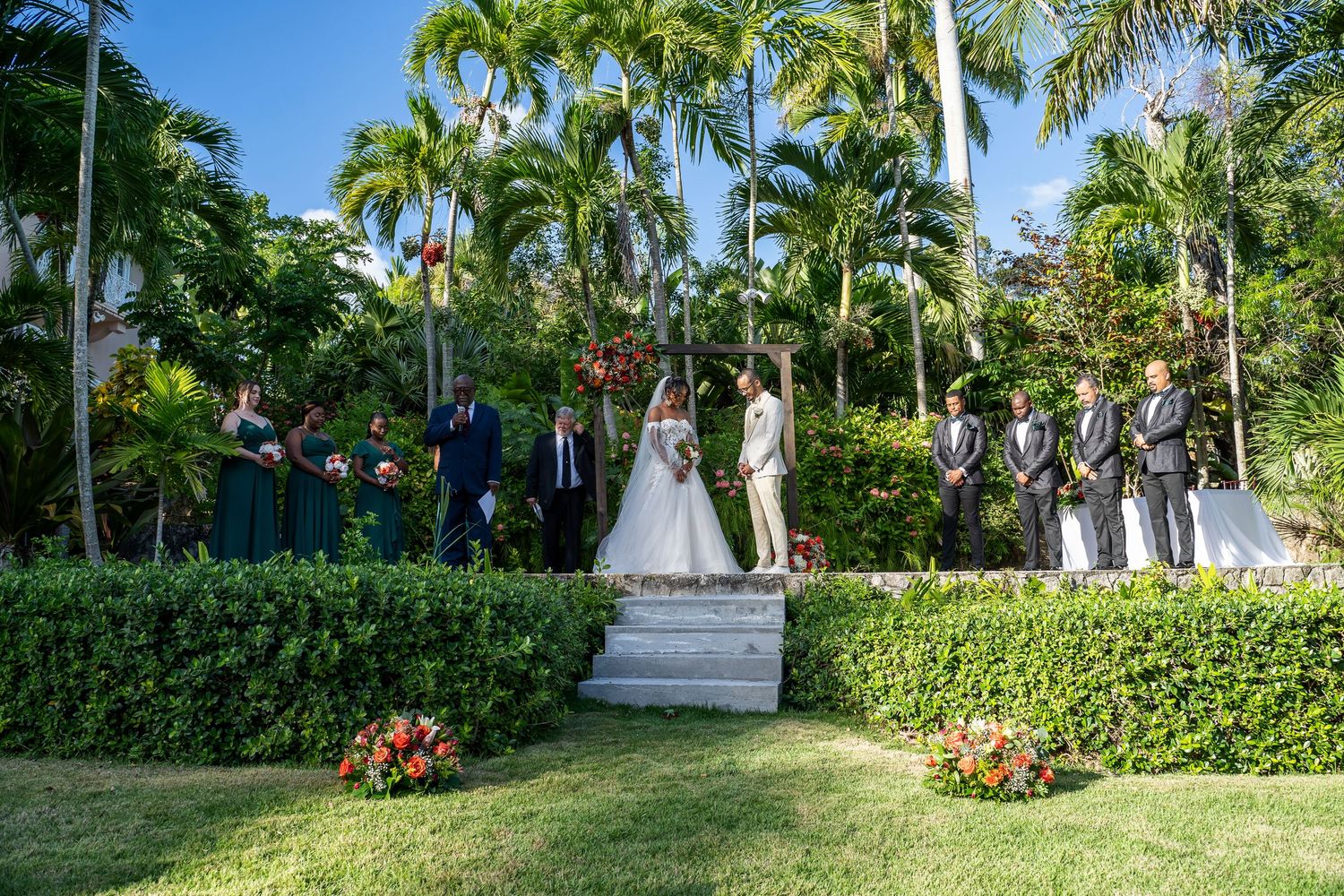 Wedding party stands on steps surrounded by tall palm trees during outdoor garden ceremony.