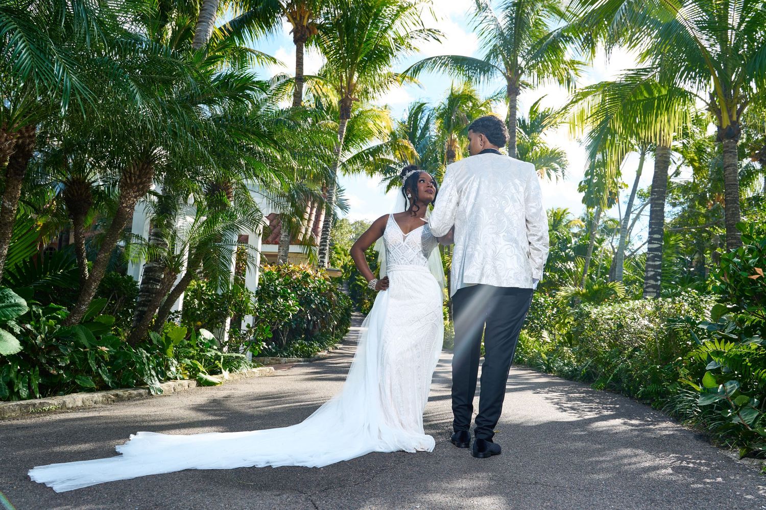 Bride and groom stroll together along a sunlit tropical path lined with tall swaying palm trees.