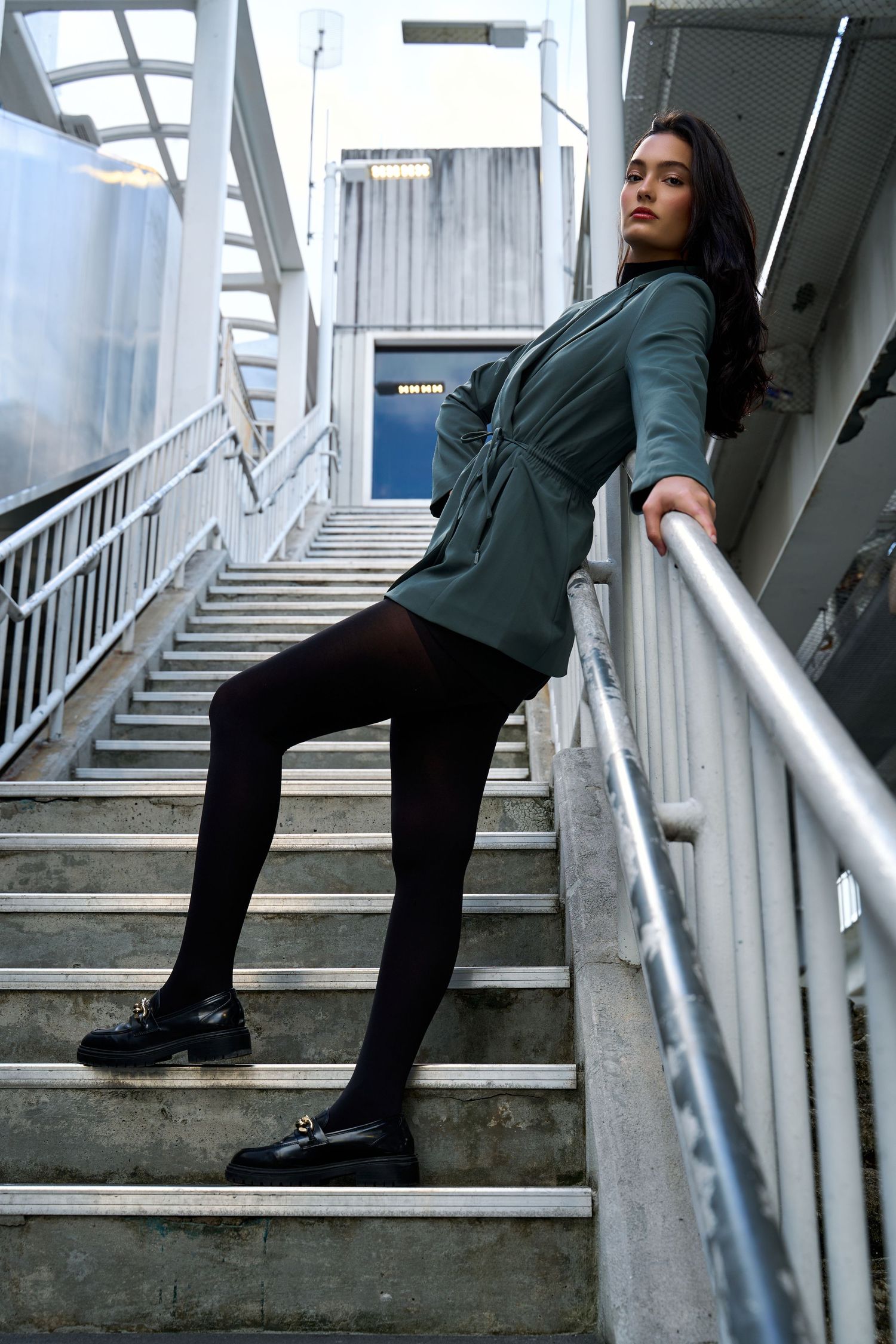 Elegant woman in teal wrap dress poses on urban staircase with black tights and loafers.