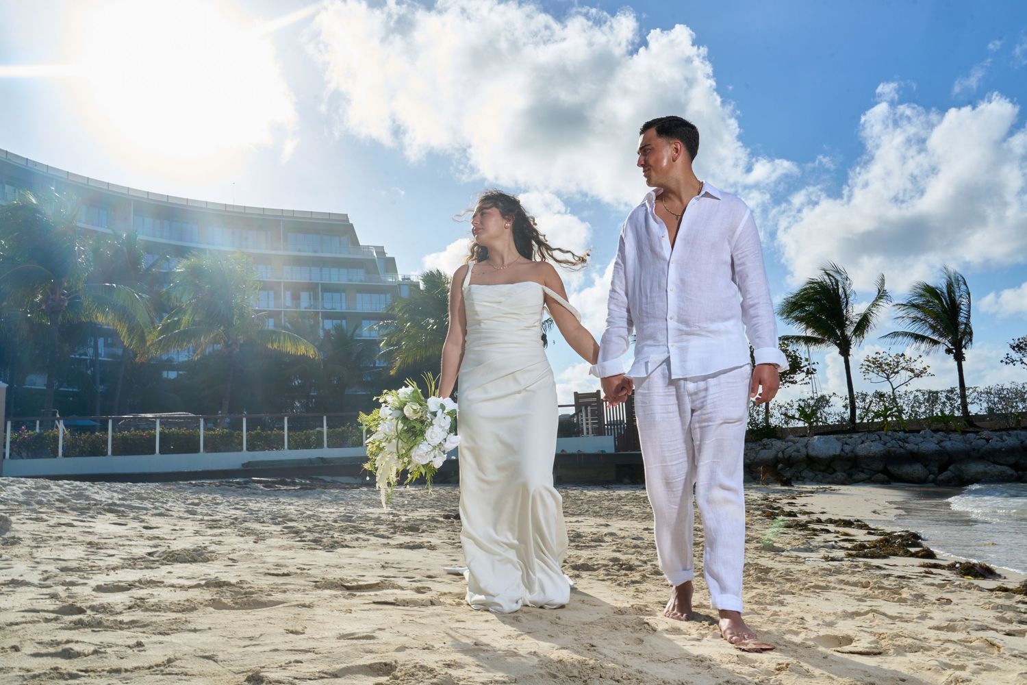Bride and groom walk hand in hand on a sunny beach with a luxury resort behind them.