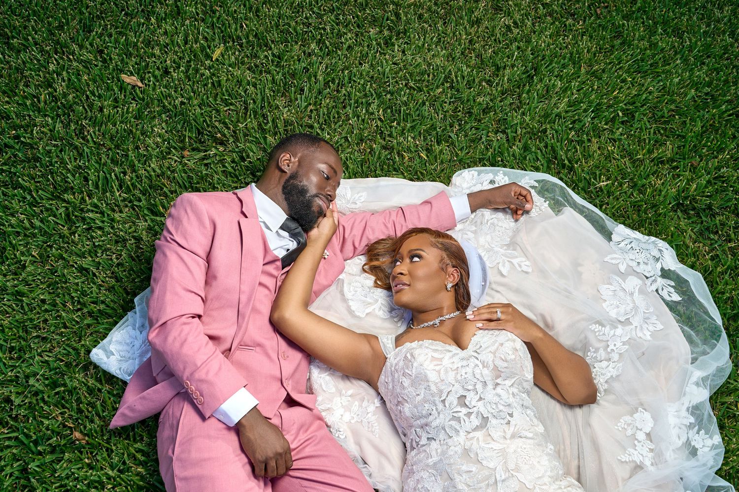 Newlywed couple lying on green grass, groom in pink suit and bride in white lace wedding dress.