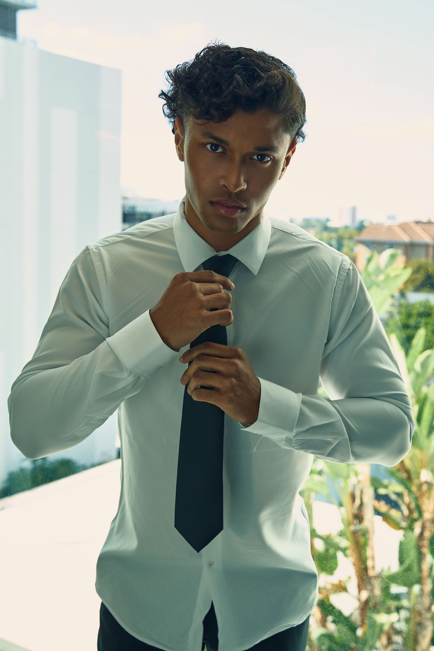 Stylish young man in white dress shirt and black tie adjusts his tie on a bright outdoor balcony.