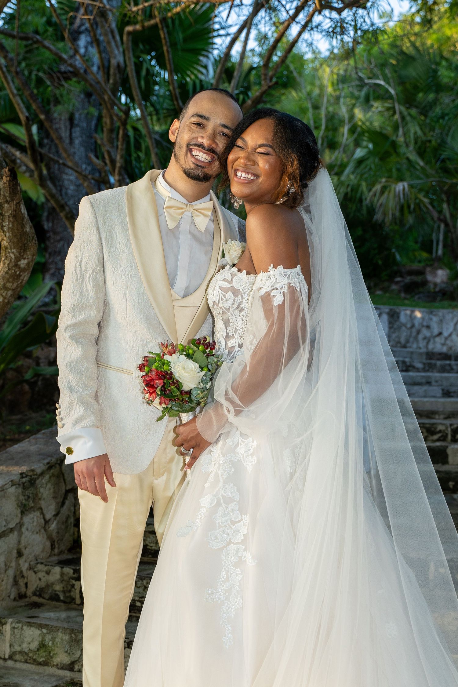 Smiling newlywed couple in cream wedding attire pose outdoors with tropical greenery and bridal bouquet.