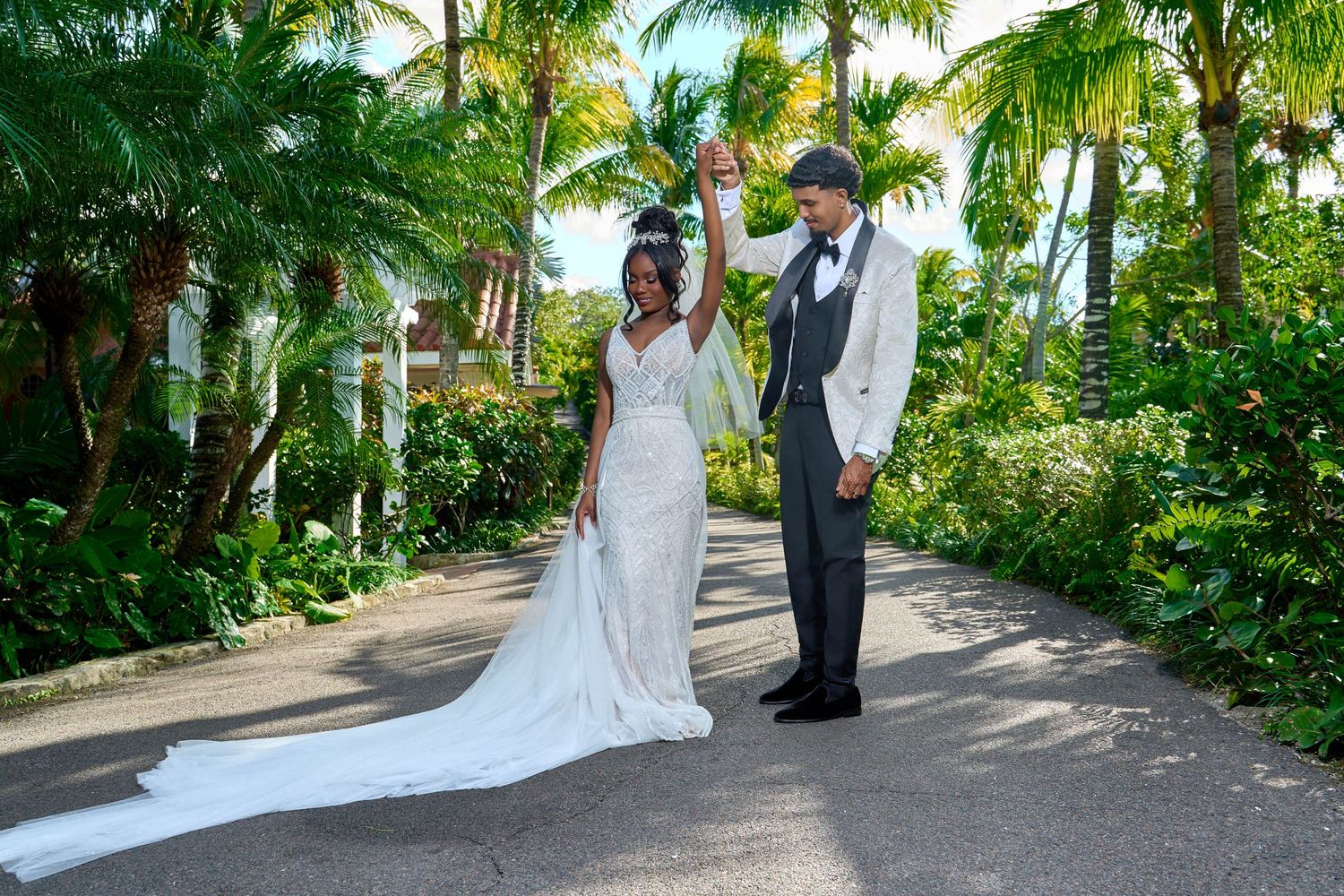 Newlywed couple celebrates with raised hands on tropical garden pathway in wedding attire.