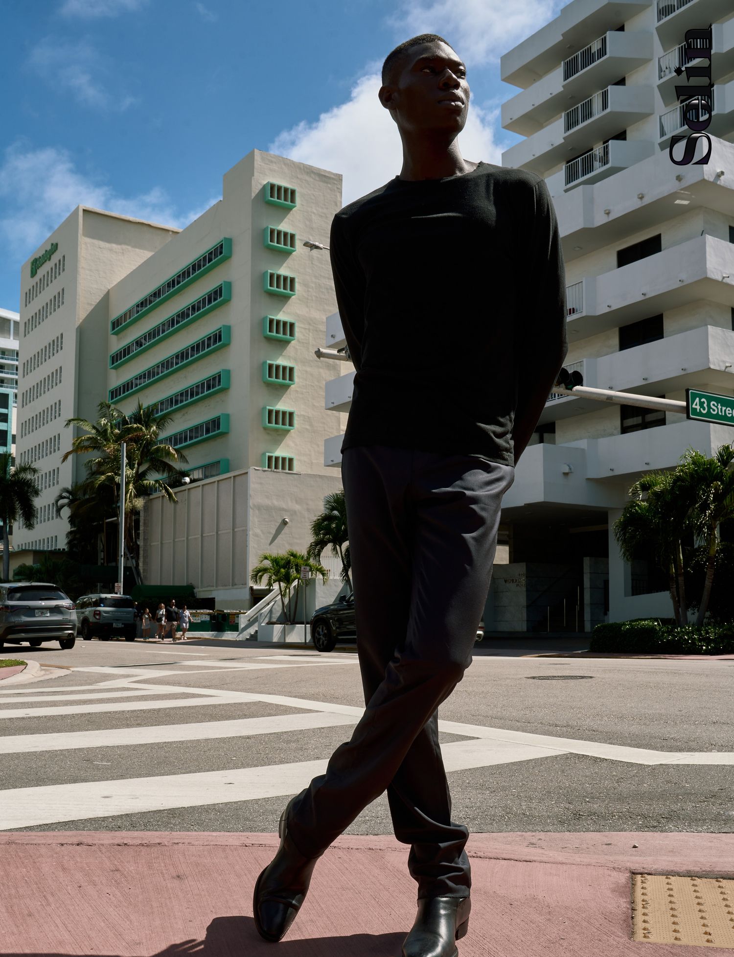 Tall man in black outfit crossing a street in Miami Beach near 43rd St with modern buildings in background.