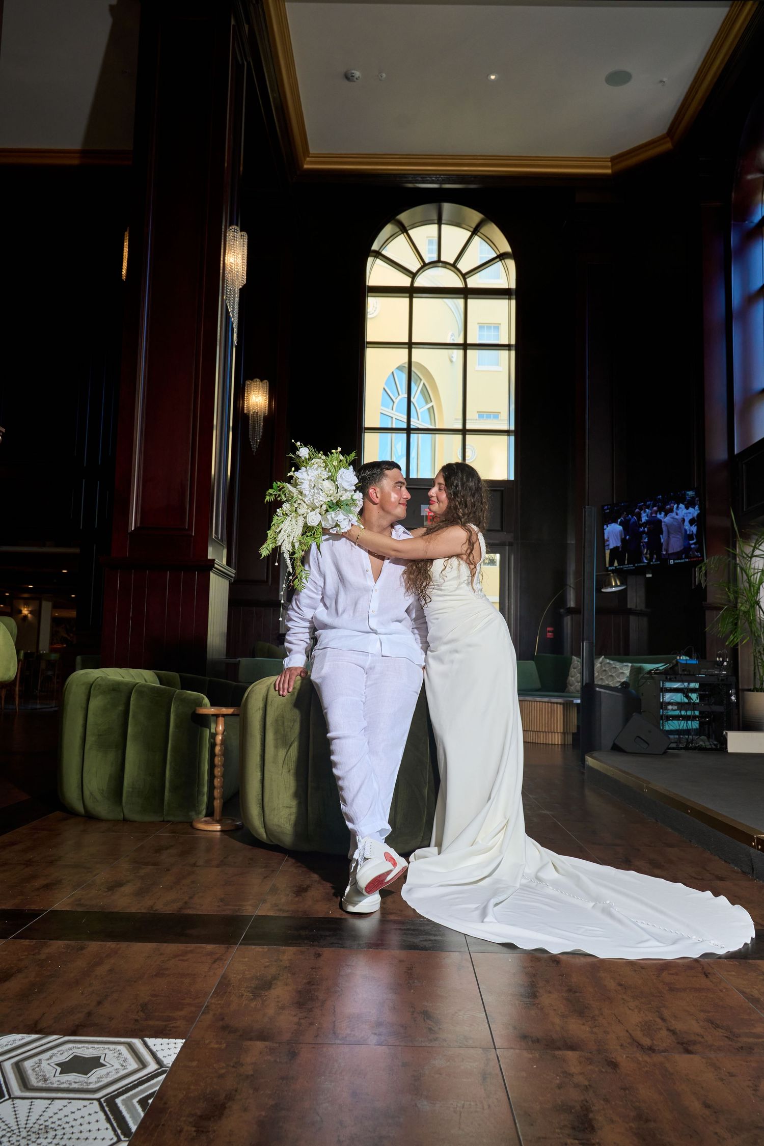 Bride and groom share a romantic moment in an elegant hotel lobby with arched windows.