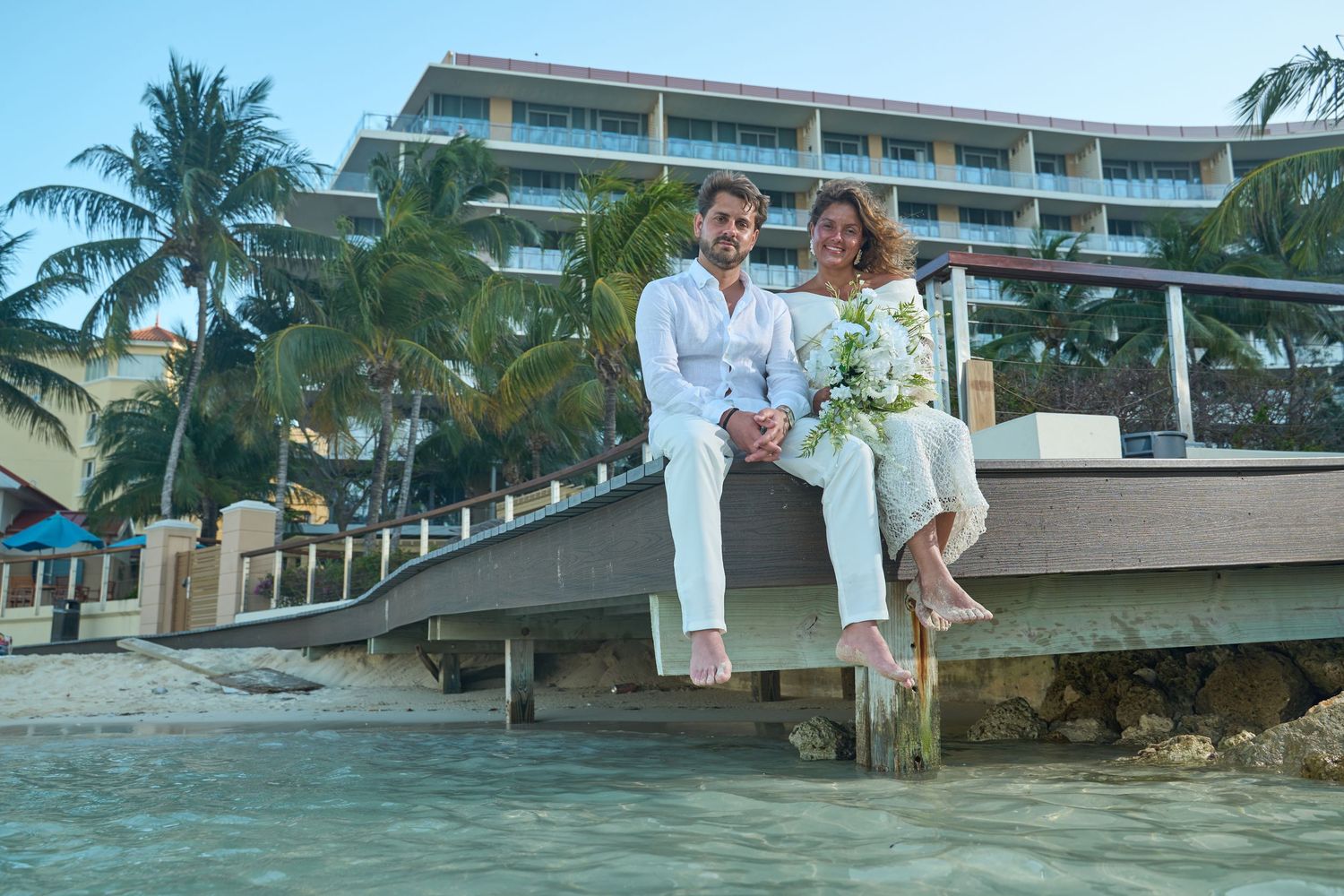 Elegant couple in white wedding attire seated on dock with tropical resort and palm trees behind.