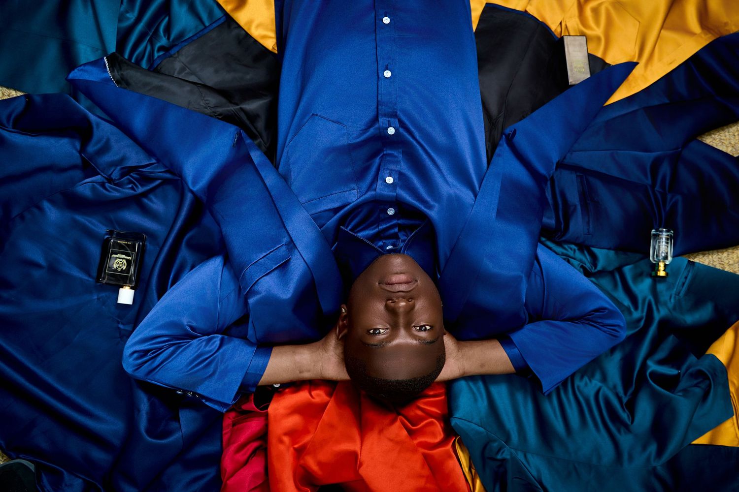 Man in blue shirt lying surrounded by colorful fabric swatches and clothing samples.