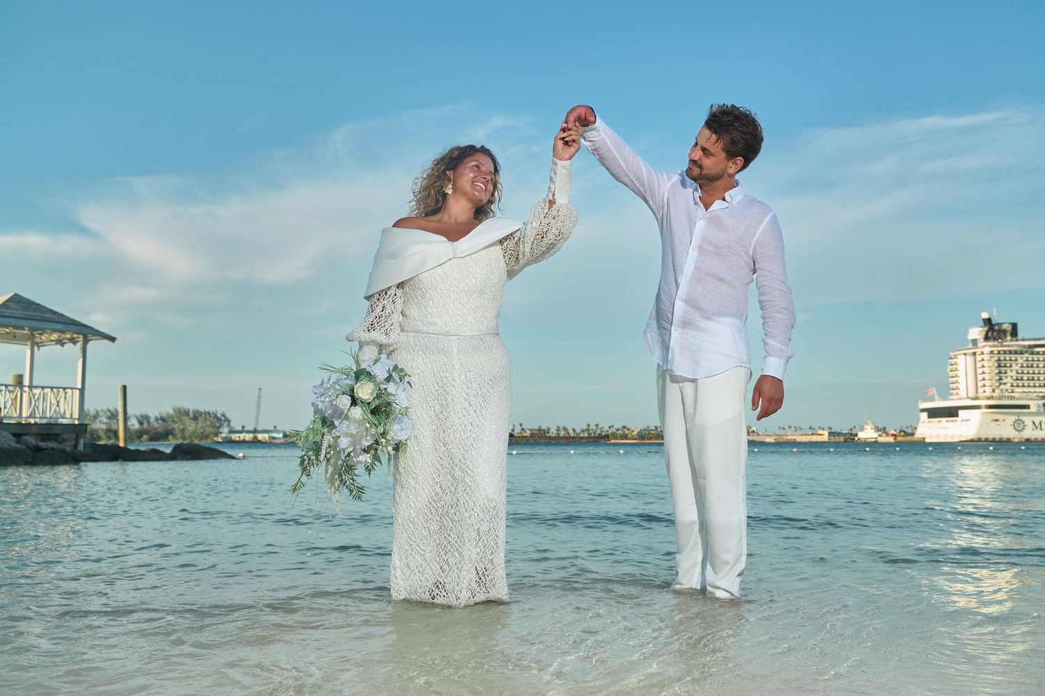 Newlywed couple dancing in shallow ocean water with harbor and cruise ship in background.