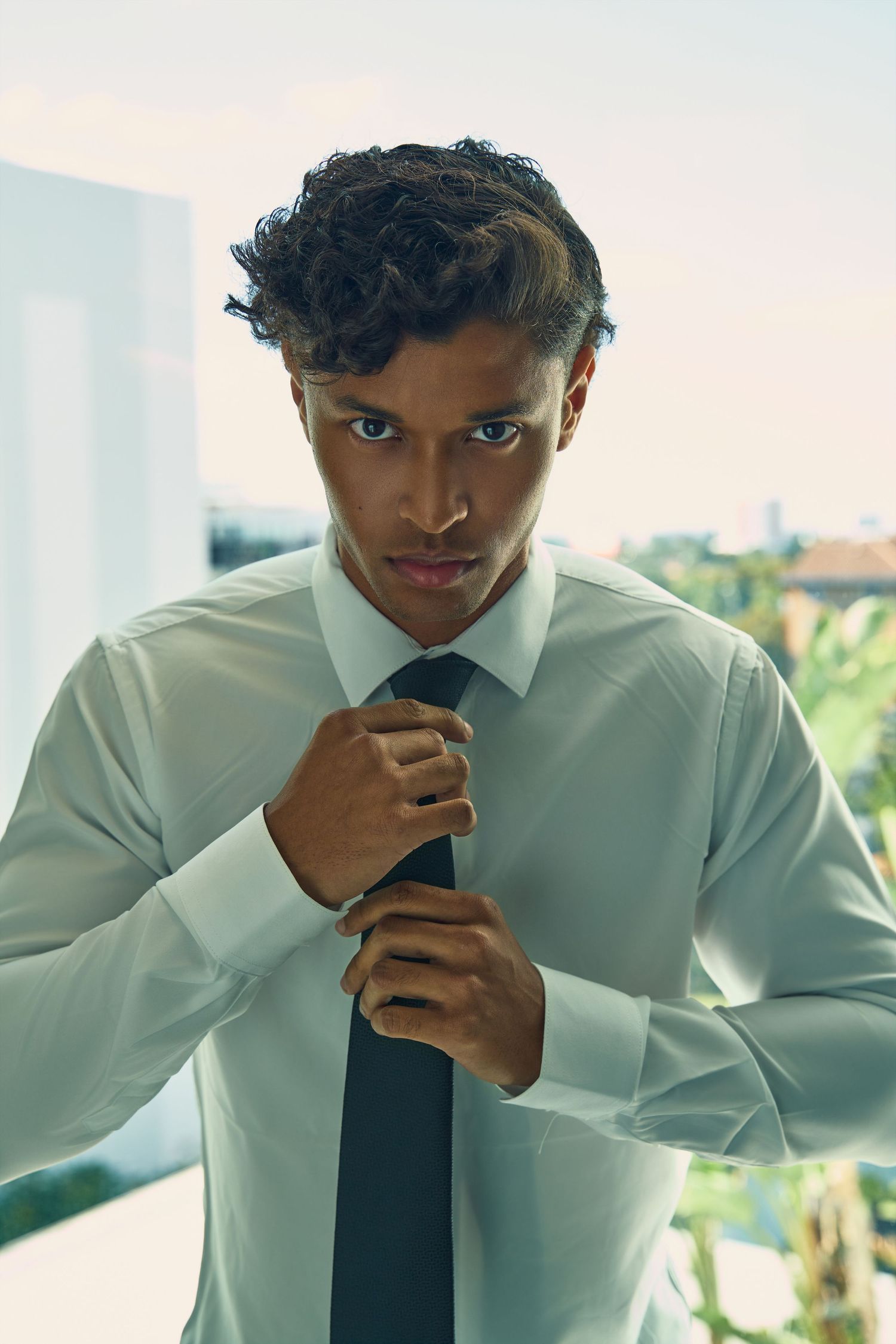 Young man in white dress shirt adjusts black tie while standing on sunlit balcony with city view.