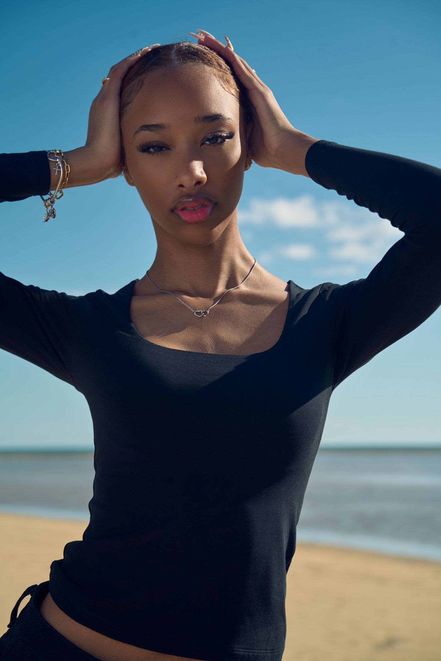 Young Black woman in black long-sleeve crop top posing on a sunny beach with ocean in the background.