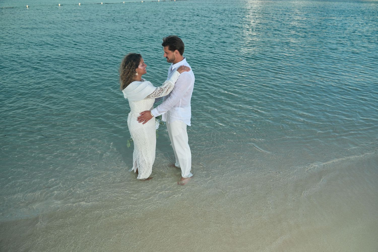 Couple dressed in white embracing romantically while standing in calm turquoise beach waters.