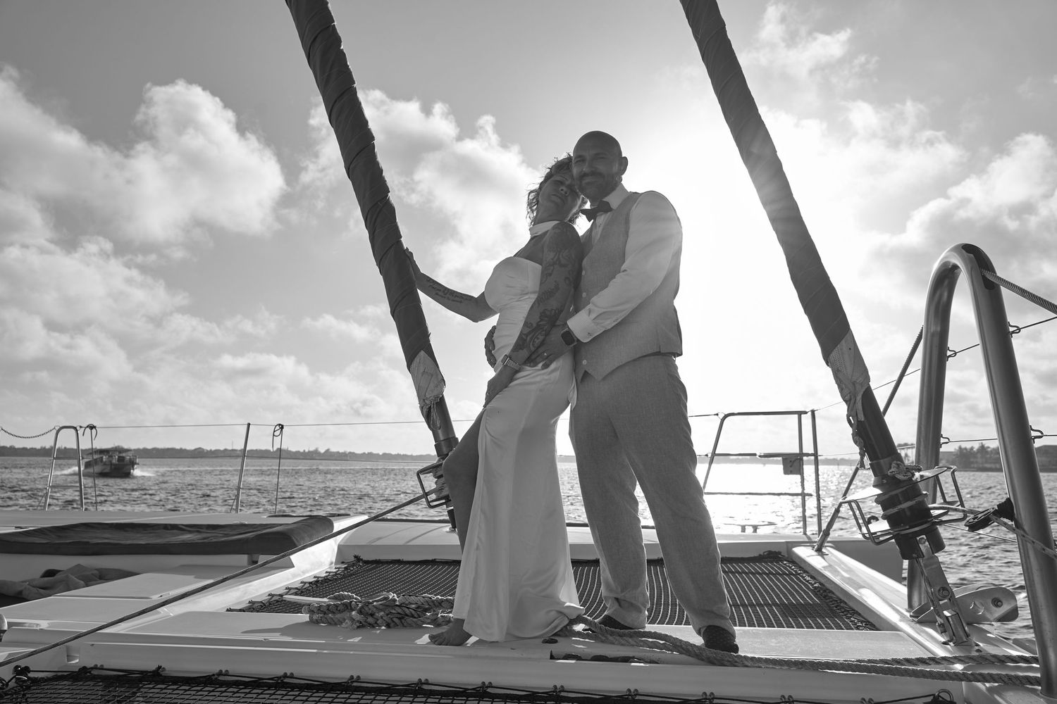 Bride and groom sharing intimate kiss on sailboat deck with sunny beach backdrop in black and white.