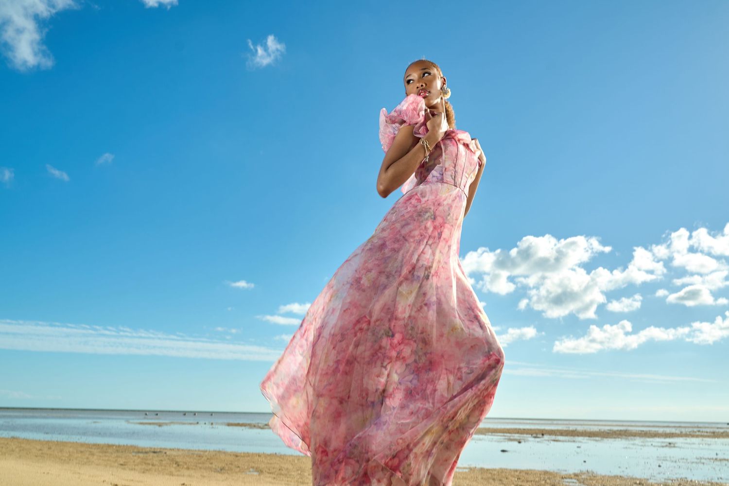 Woman in flowing pink floral maxi dress standing on a beach with blue sky and ocean behind her.