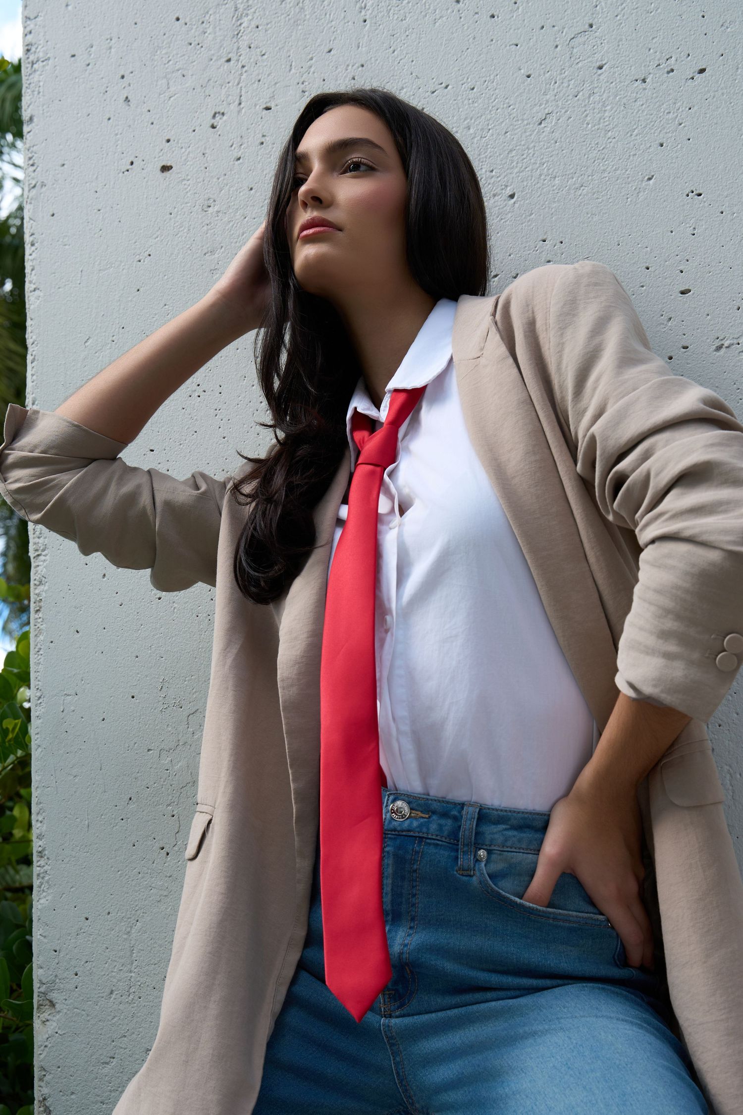 Stylish woman in beige blazer, white shirt, red tie, and jeans leans against white wall in urban setting.