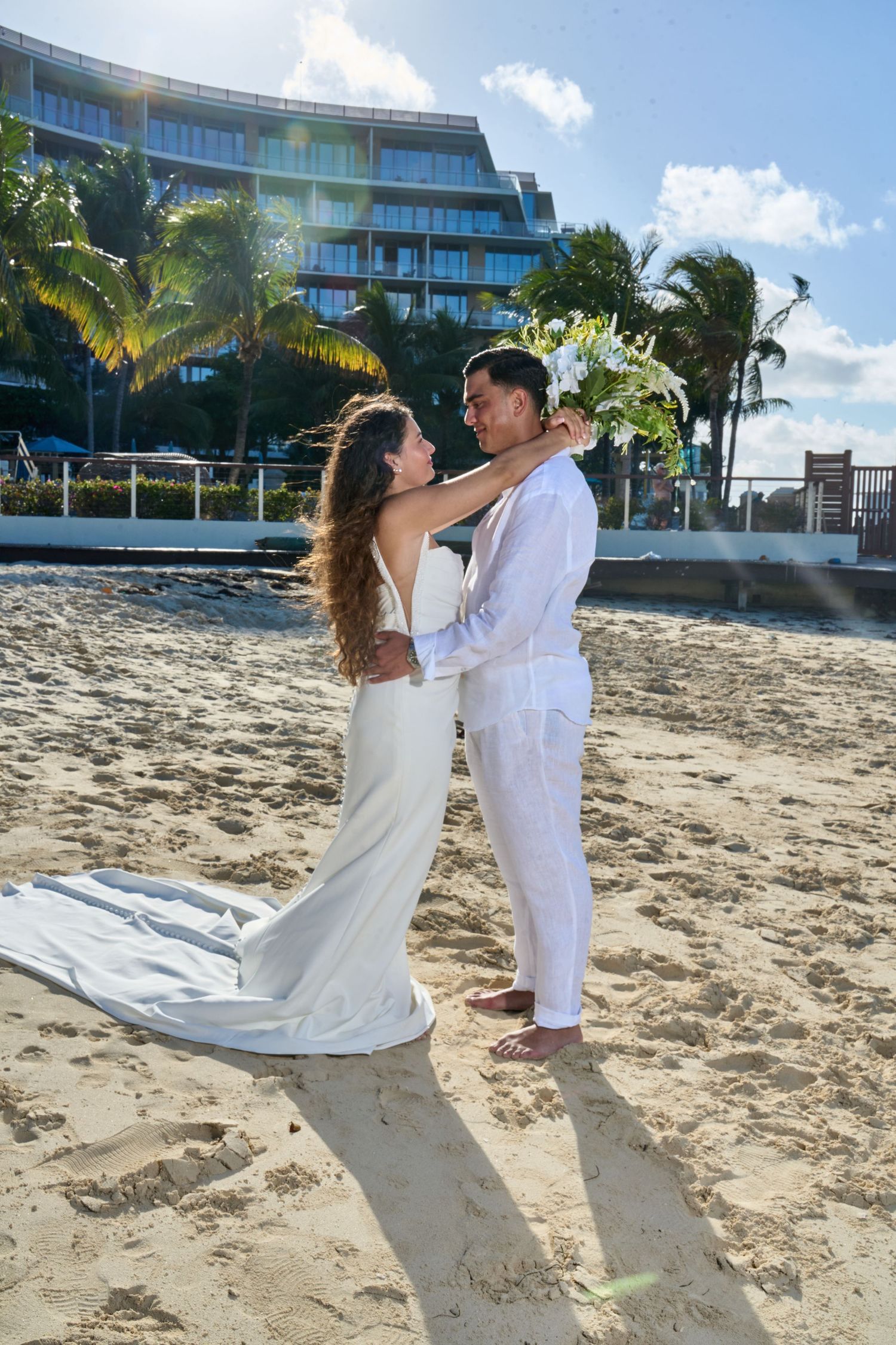 Bride and groom embrace on sandy beach with palm trees and modern resort building backdrop.