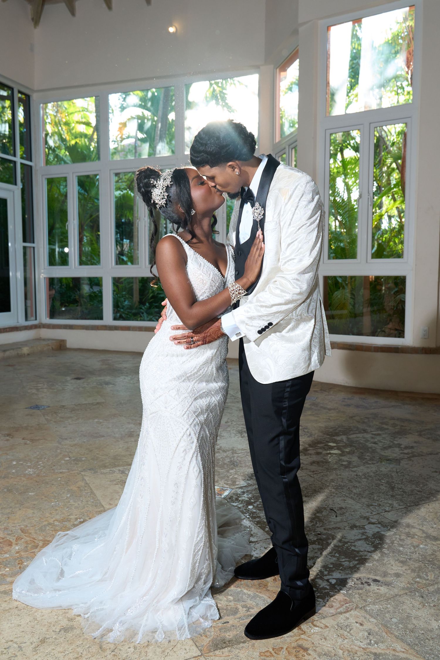 Newlywed couple sharing a romantic kiss indoors, bride in beaded gown and groom in white tuxedo jacket.