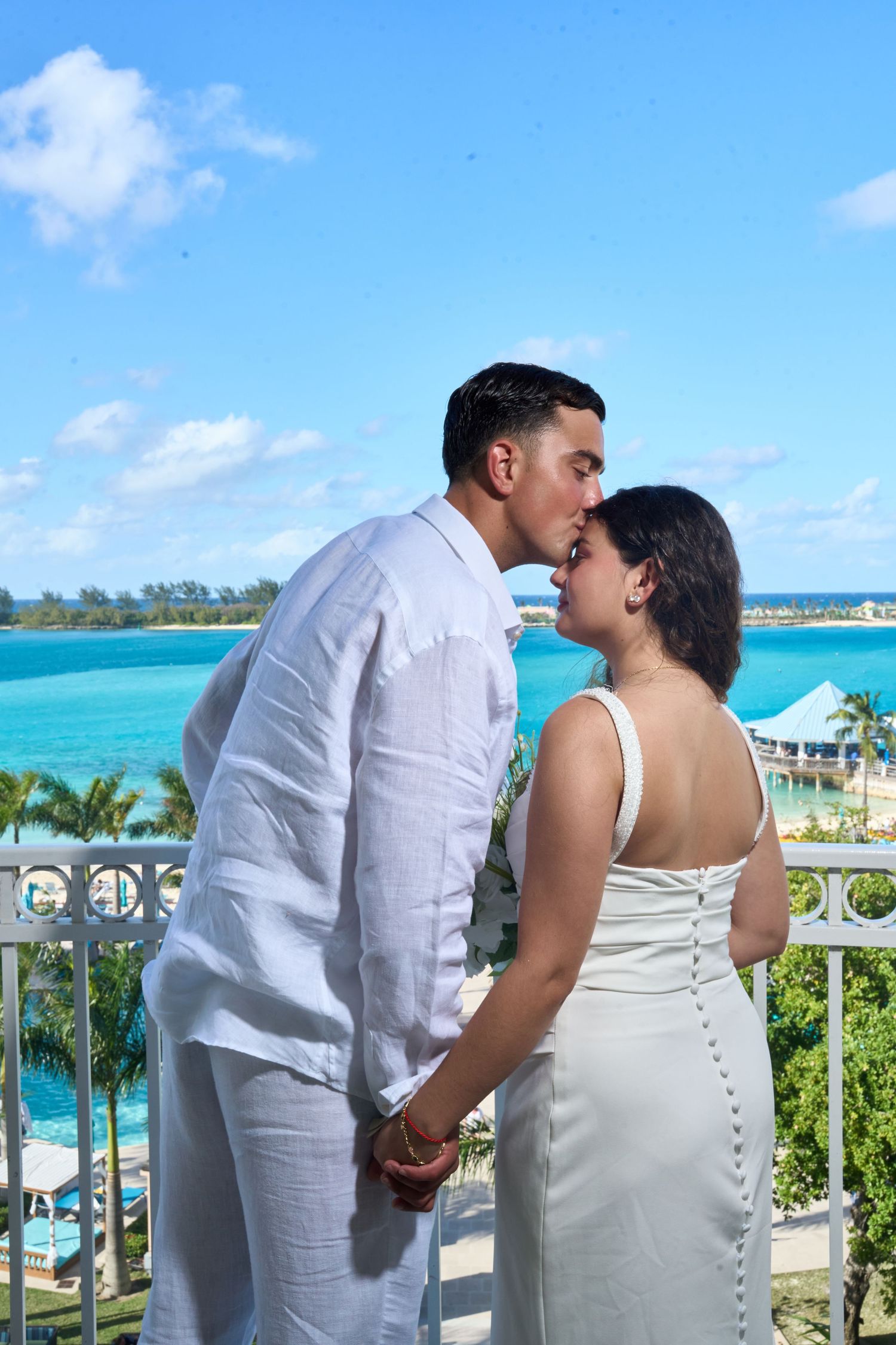 Groom kisses bride on a balcony with breathtaking turquoise ocean and tropical scenery behind them.