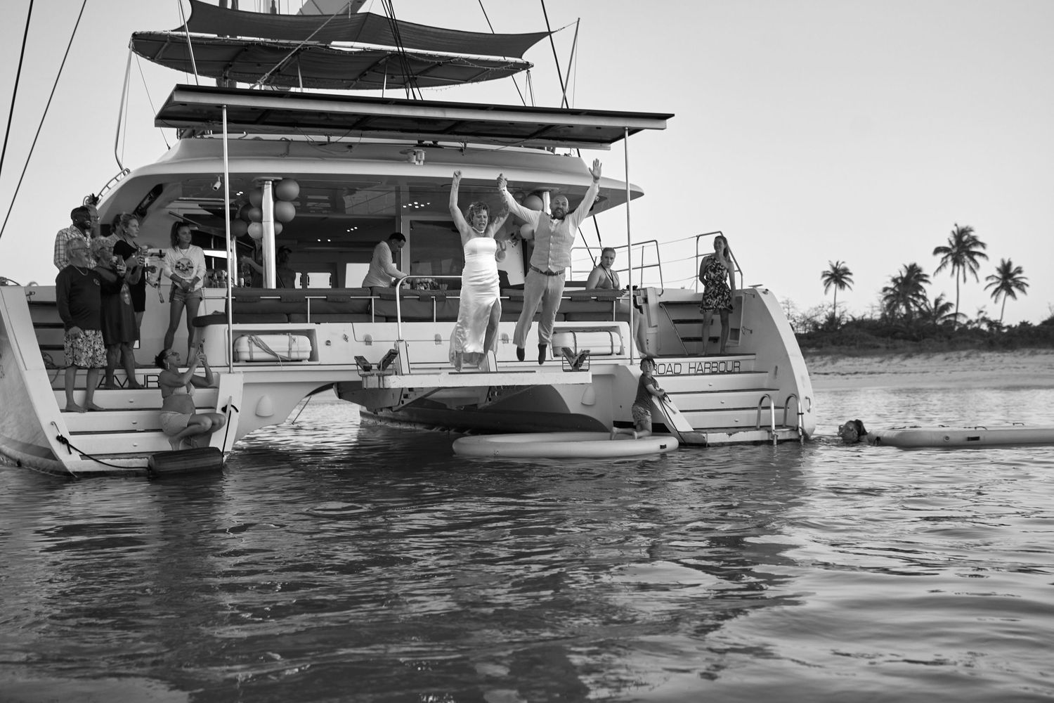 Group of guests celebrating on catamaran yacht deck in black and white tropical setting.