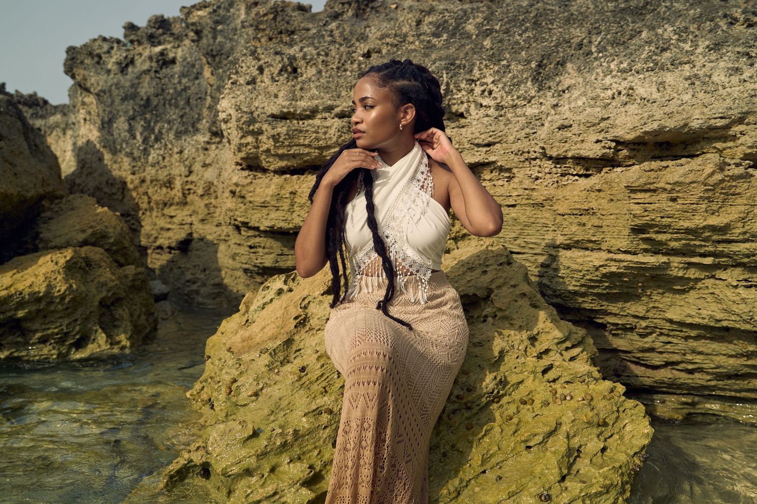 Woman in fitted crochet halter dress posing against rugged rocky cliffs near the ocean.