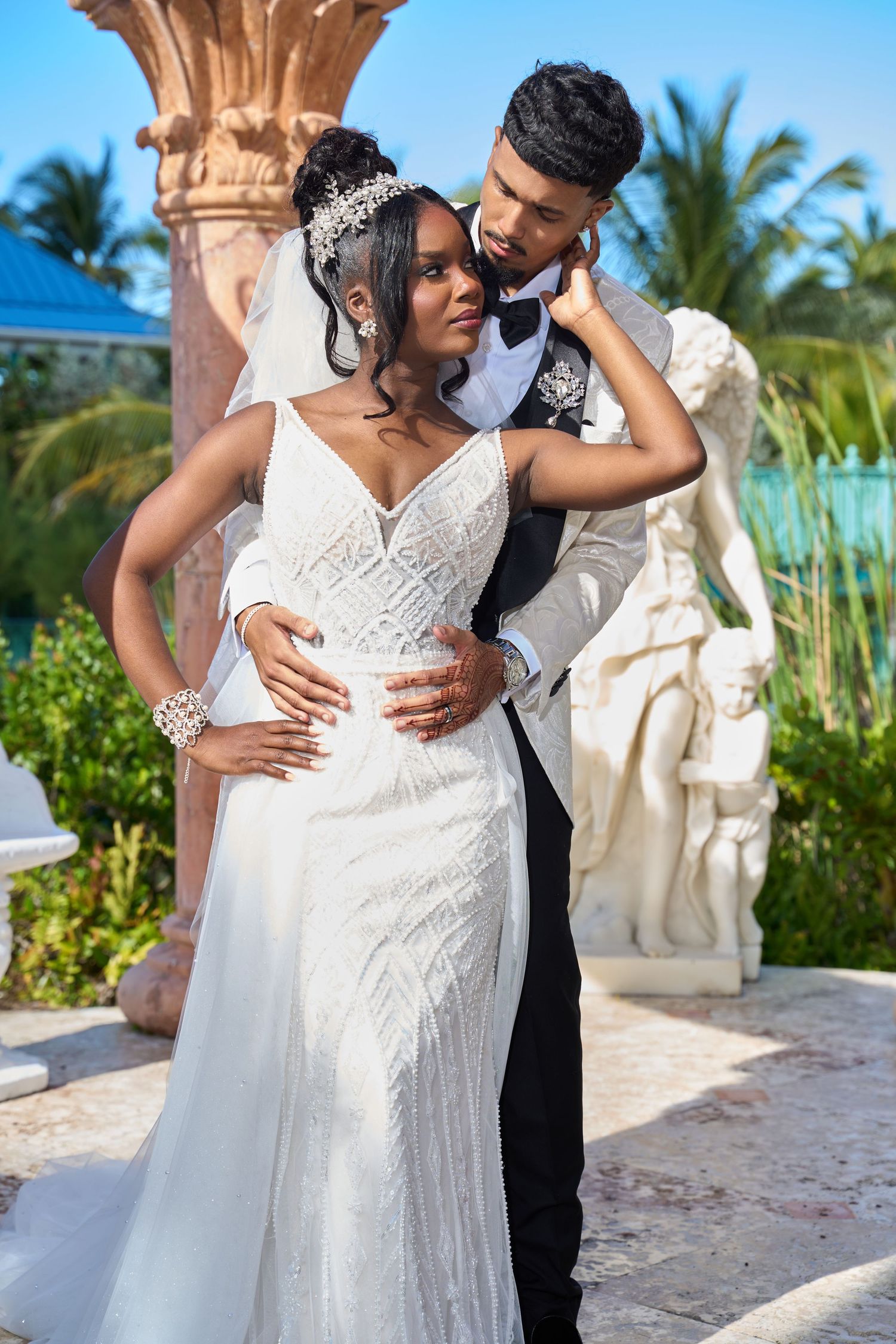 Bride in beaded gown and groom in white tuxedo embrace tenderly near classical statues at outdoor venue.