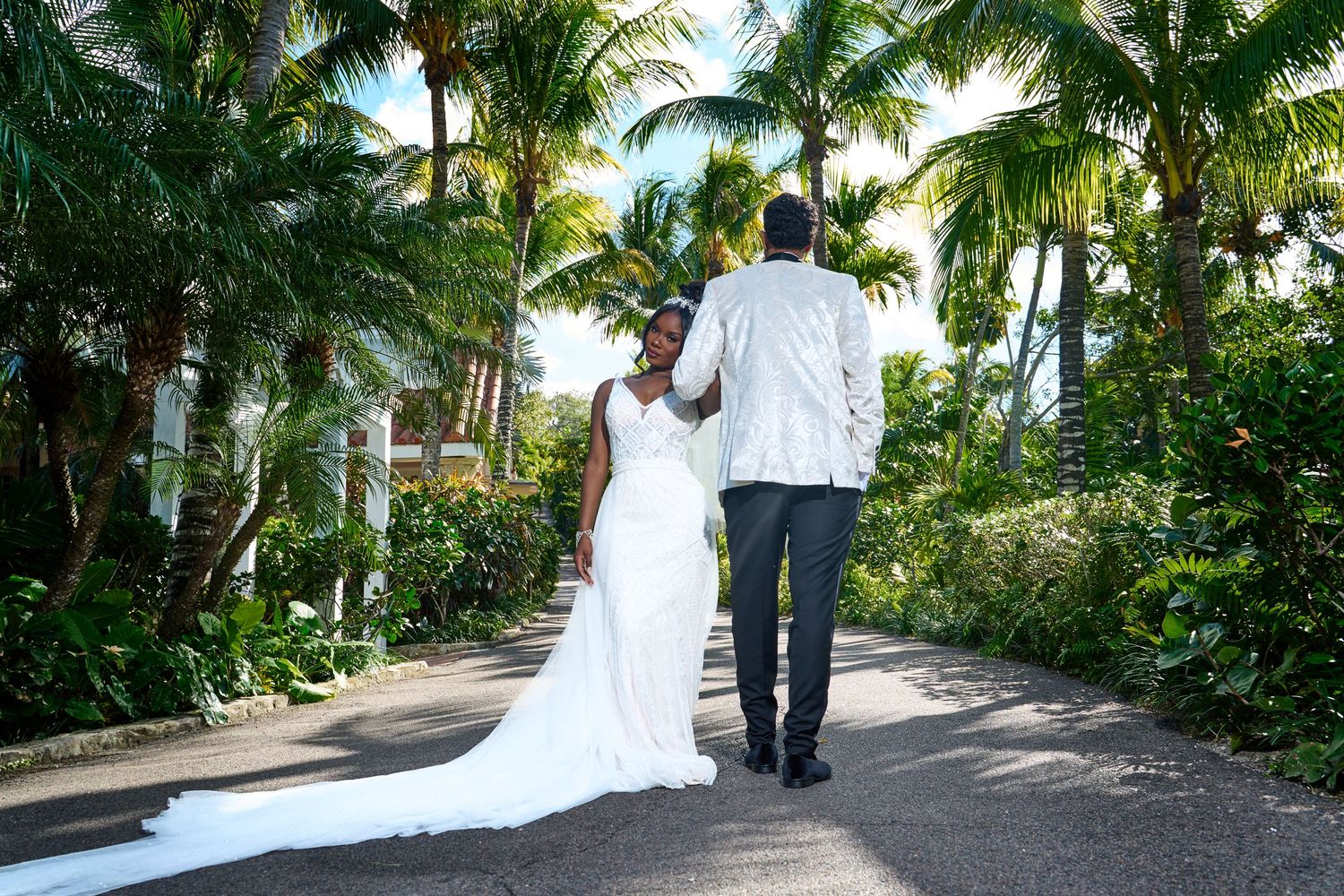 Newlywed couple walks hand-in-hand down a lush tropical palm-lined pathway after their wedding ceremony.