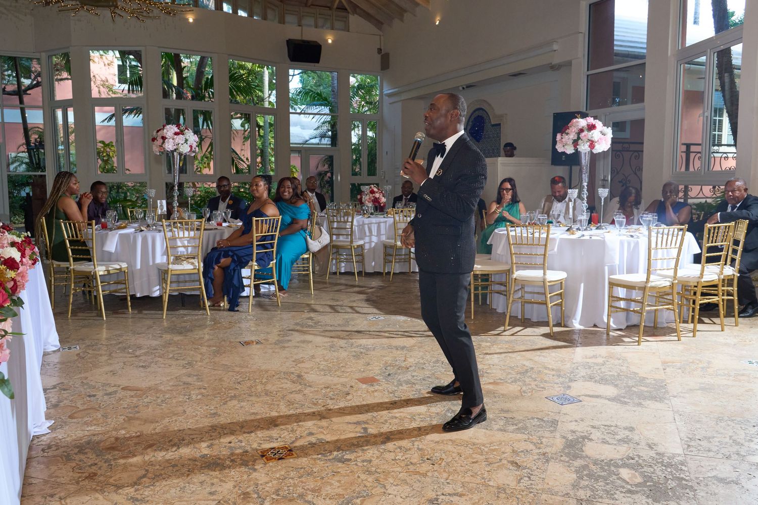 Man in tuxedo giving speech at elegant wedding reception with gold chairs and floral decorations.