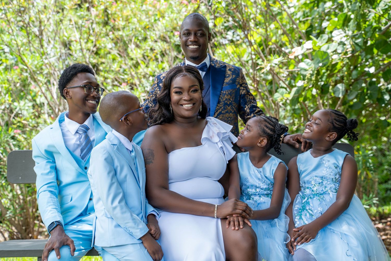 Joyful family wedding portrait with bride, groom, and three children in coordinating blue outfits outdoors.