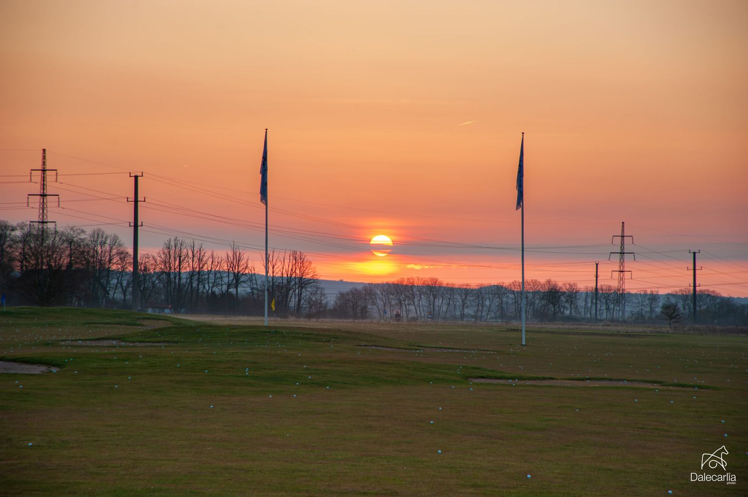Stunning sunset over a golf course with flags and power lines silhouetted against an orange sky.