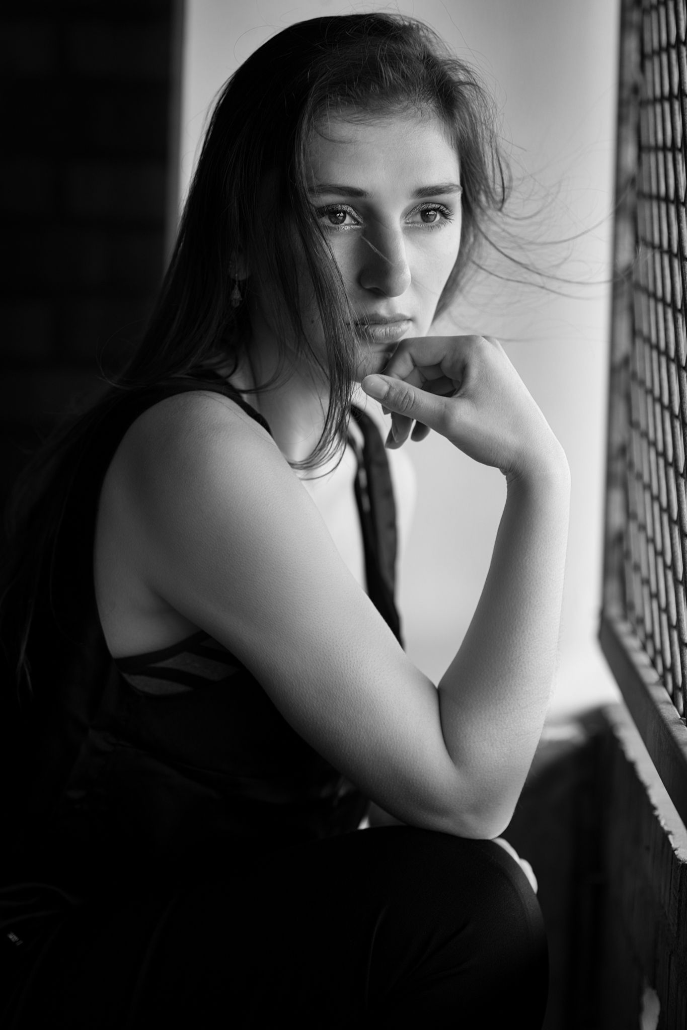 Black and white portrait of a young woman with long hair, resting her chin on her hand near a window.