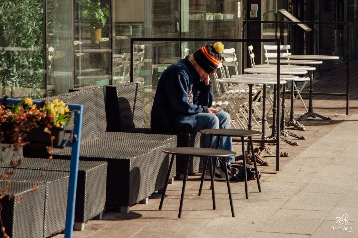 Person in colorful hat sitting alone at outdoor cafe tables on urban sidewalk.