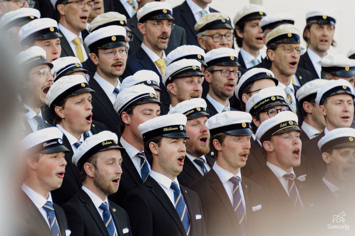 Large male choir wearing navy uniforms and white caps performing outdoors at a formal event.