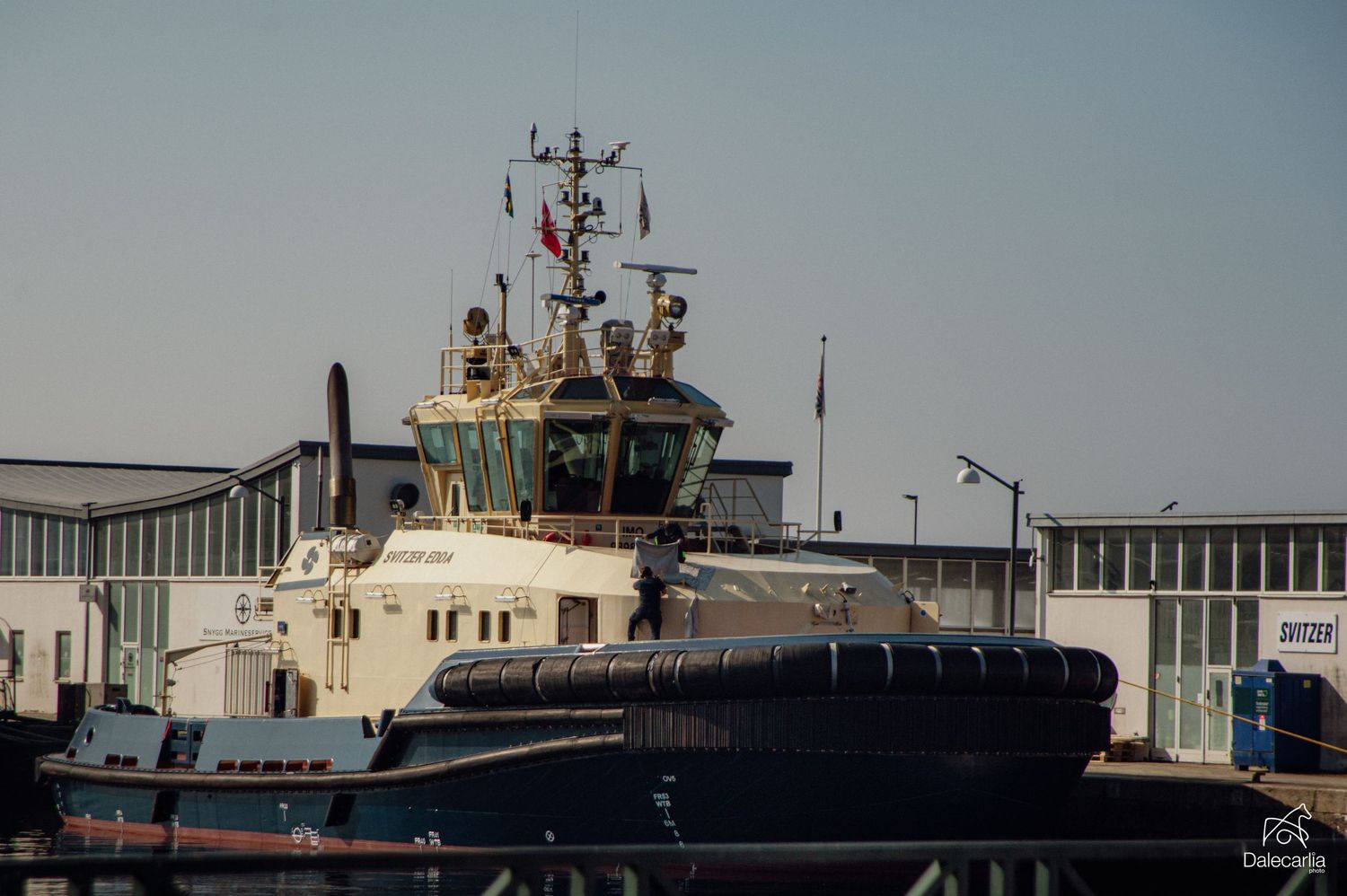 A tugboat docked at an industrial harbor under a clear sky, featuring a white hull, radar equipment, and a Turkish flag.