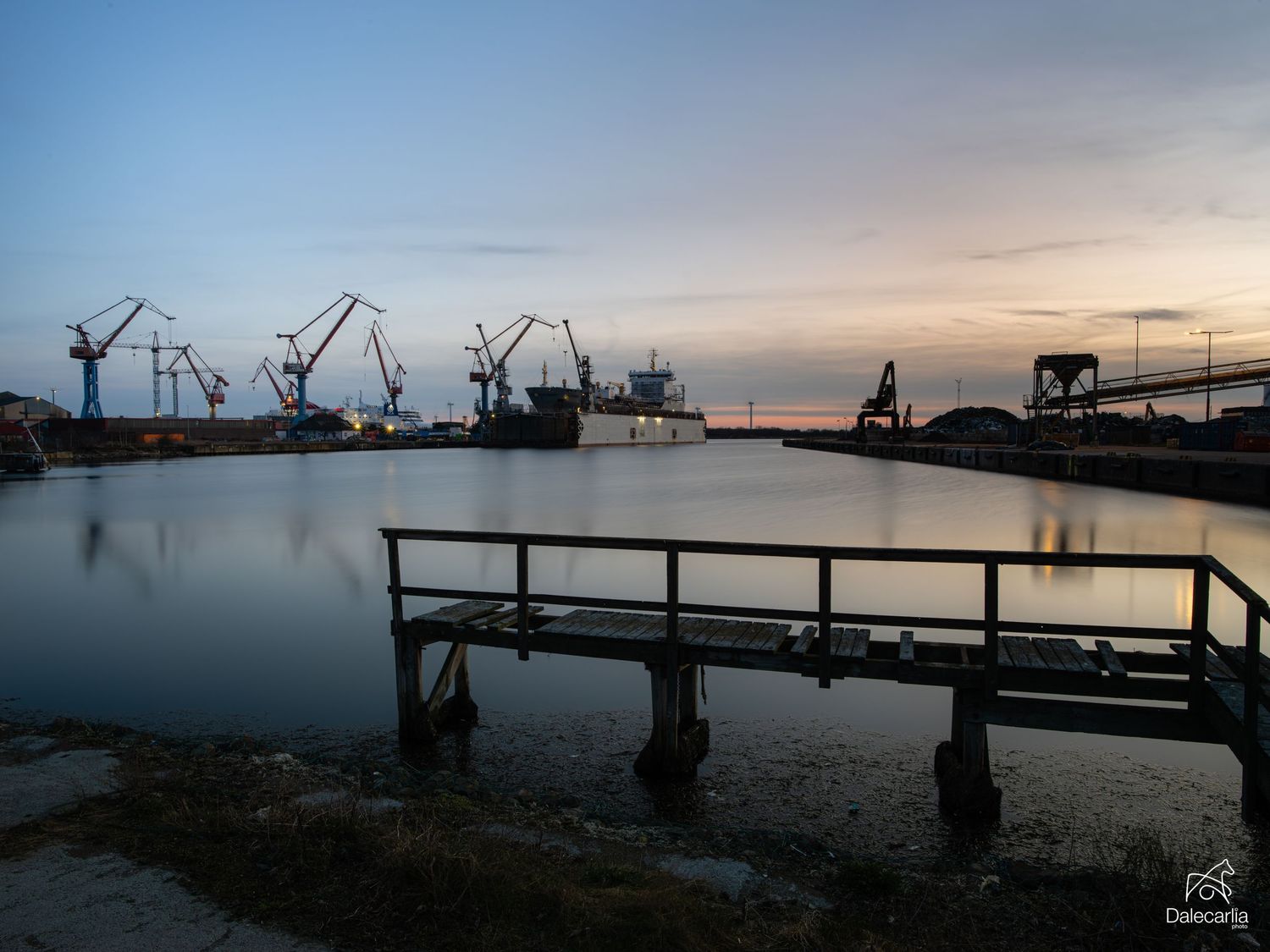 Industrial harbor at dusk with cranes and ships reflected in calm water, metal dock in foreground under colorful sunset sky.