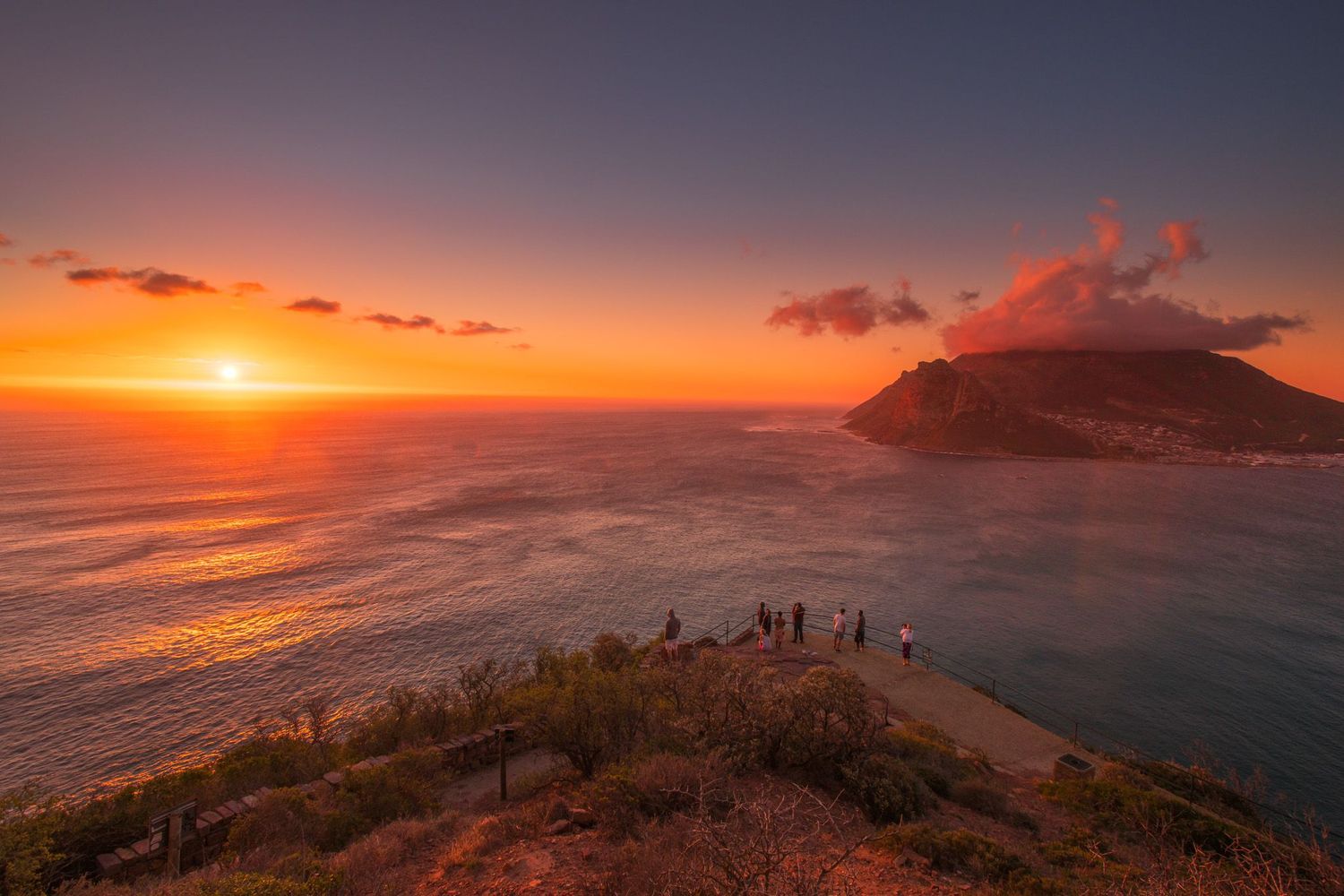 Stunning sunset over the ocean with silhouetted figures on a rocky cliff overlooking a coastal mountain landscape.