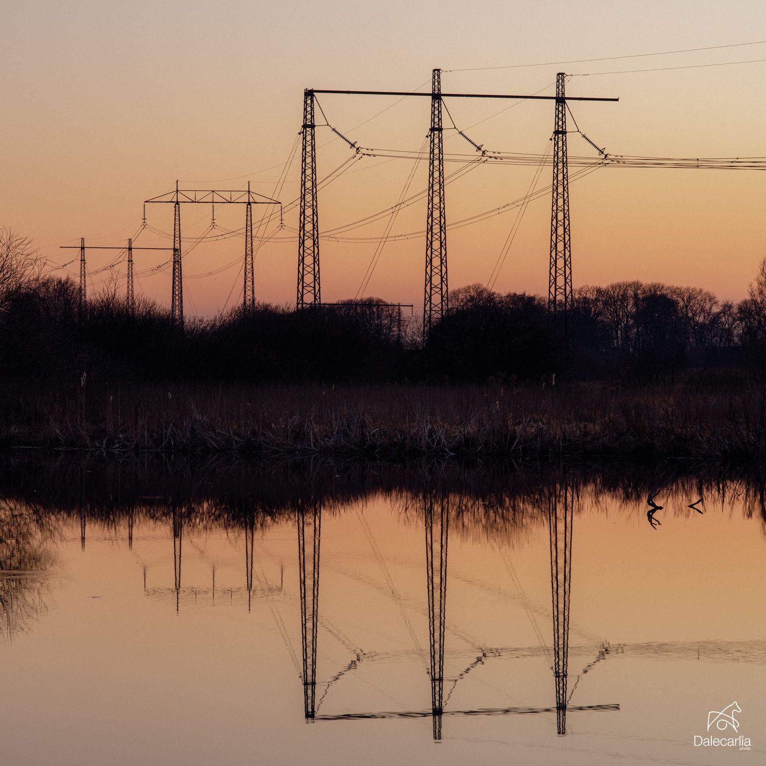 Power line towers reflected in calm water at sunset with silhouetted trees and warm orange sky.