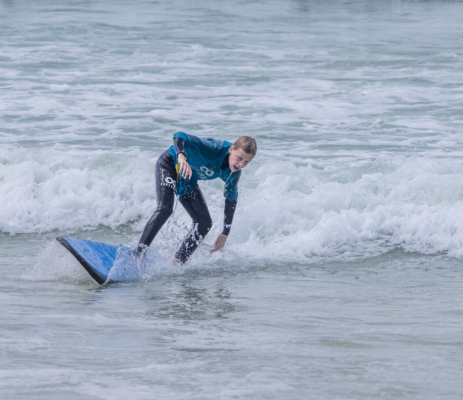 Young surfer in a black wetsuit riding a blue surfboard on a small wave in the ocean.