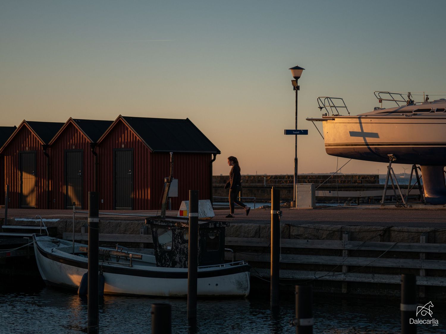 Person walking on a wooden dock at sunset with red fishing huts and a docked sailboat in a peaceful harbor.