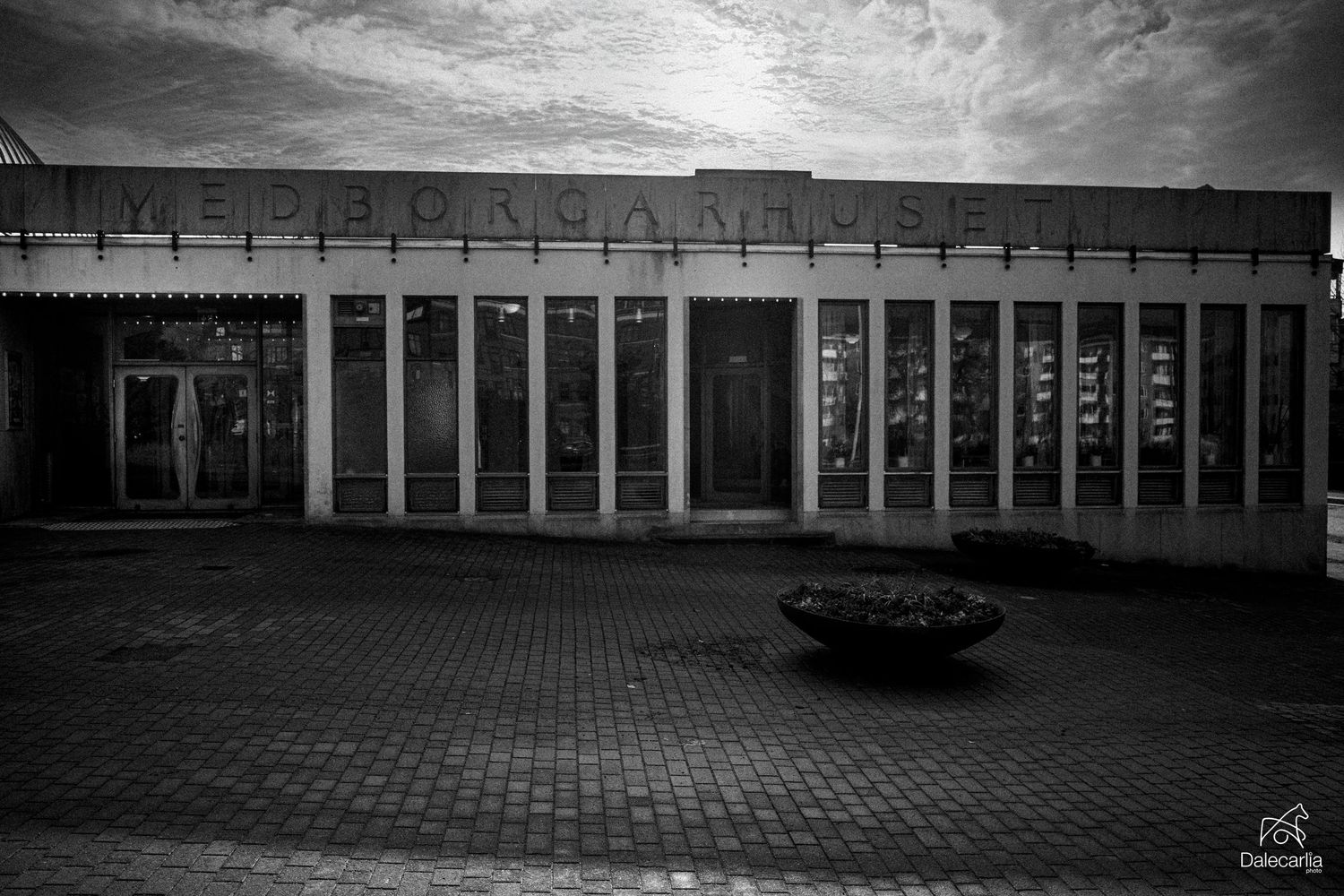 A vintage car parked in front of a modernist building with columns under dramatic cloudy skies in black and white.
